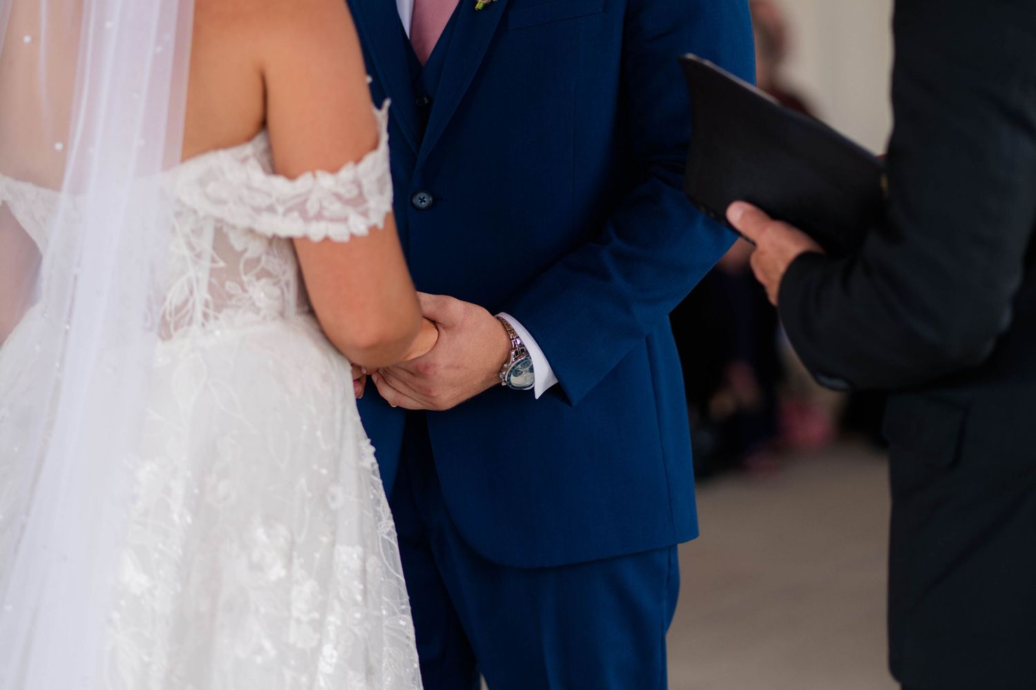 Bride in lace off-shoulder gown holds hands with groom in navy suit during wedding ceremony.