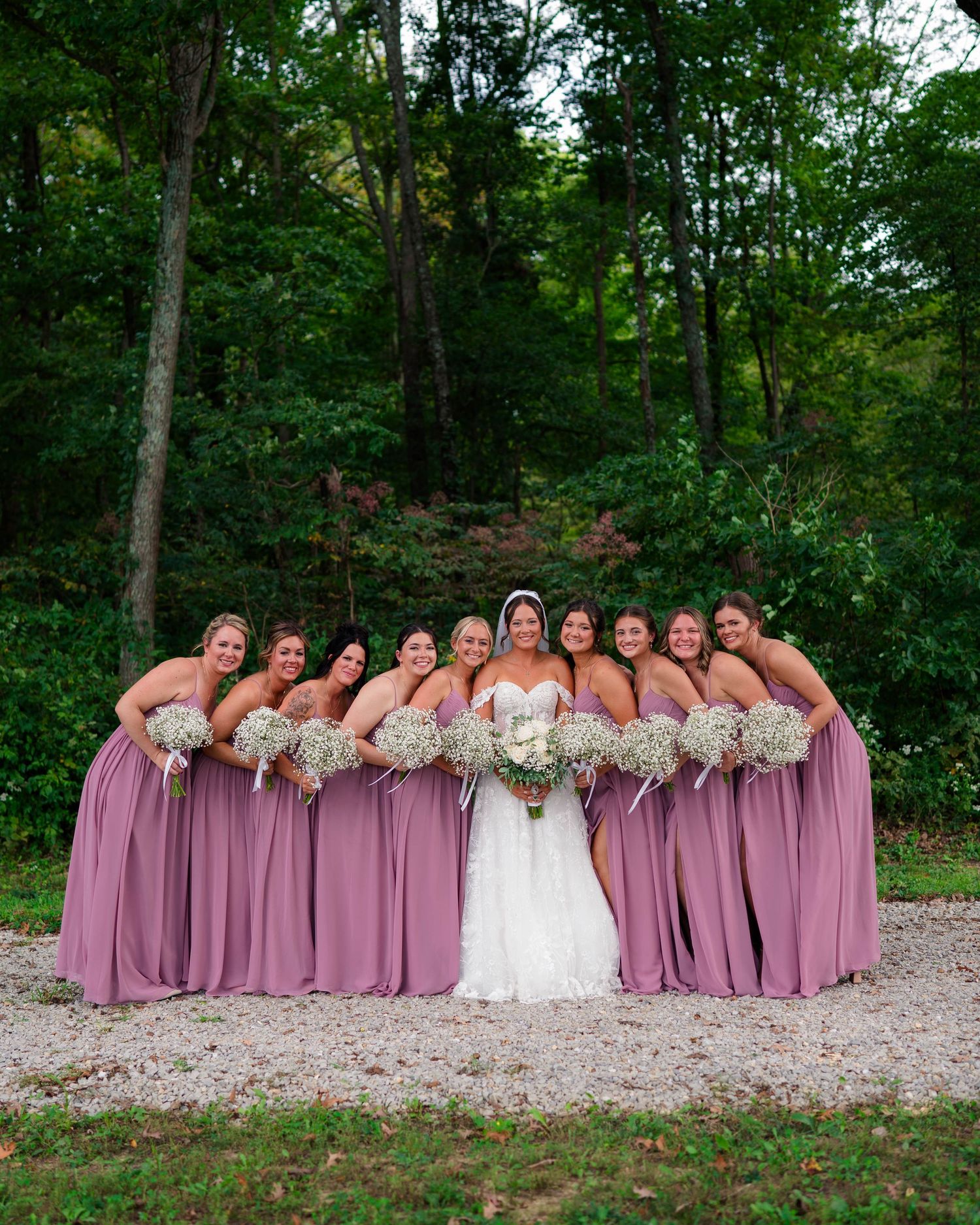 Bride in white gown poses with nine bridesmaids in mauve dresses holding baby's breath bouquets outdoors.