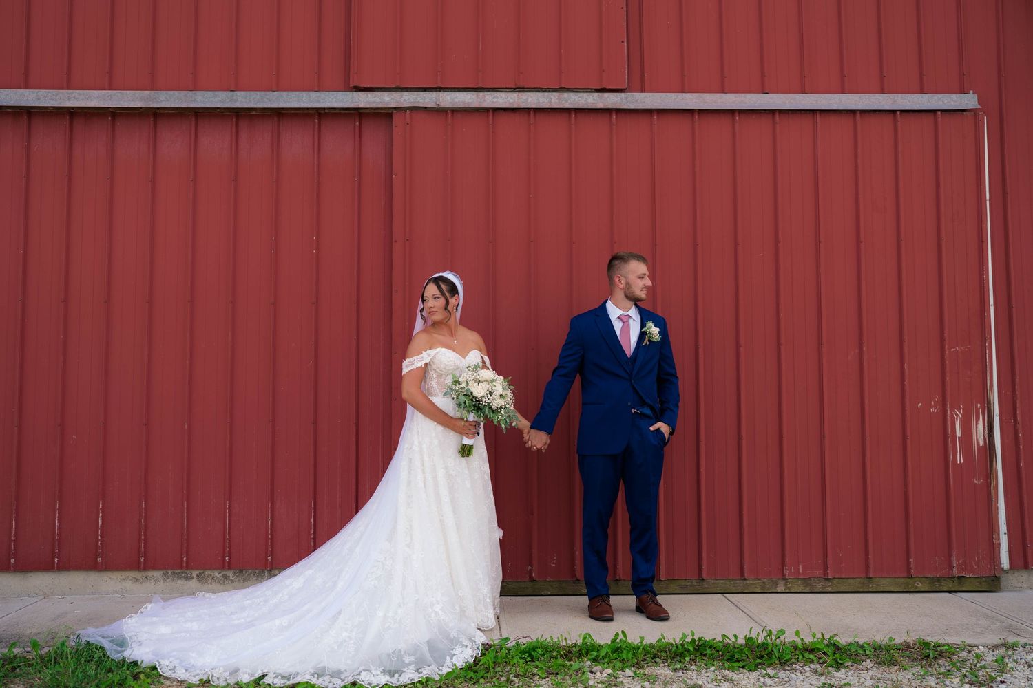 Bride in white gown and groom in blue suit hold hands posing before a red barn wall.