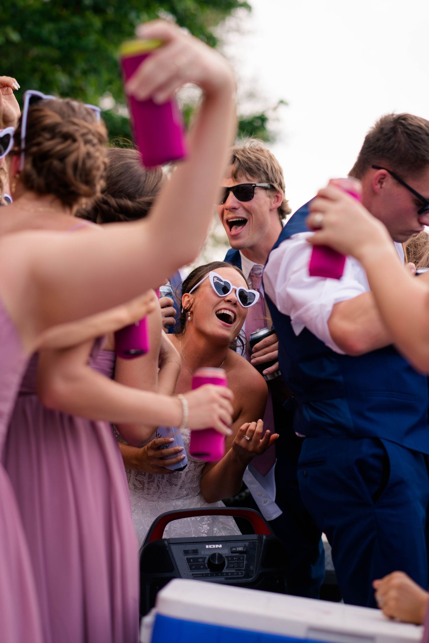 Bride and wedding guests laugh and celebrate with pink cups at outdoor reception party.