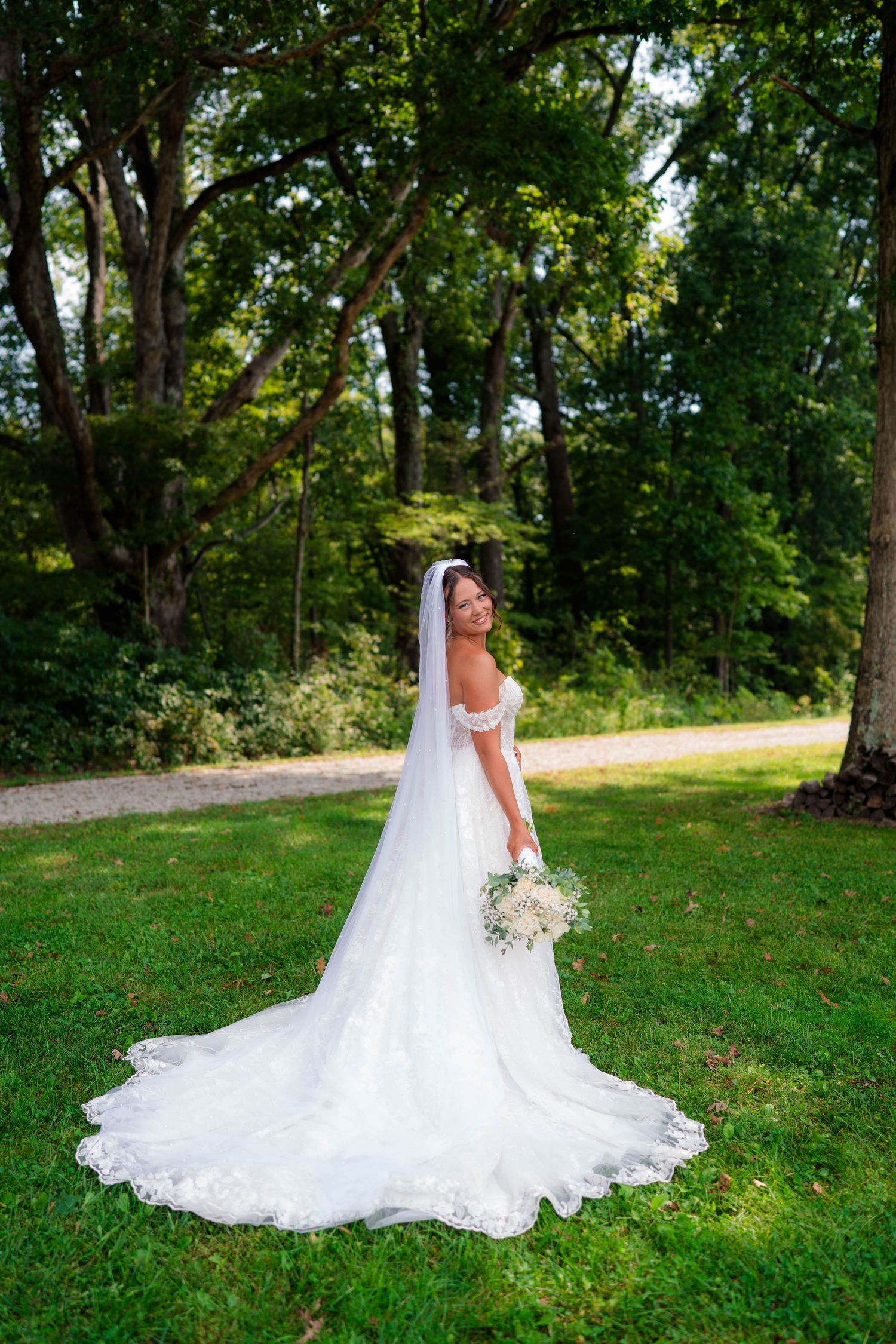 Elegant bride in off-shoulder lace wedding gown with cathedral veil stands on green lawn surrounded by trees.
