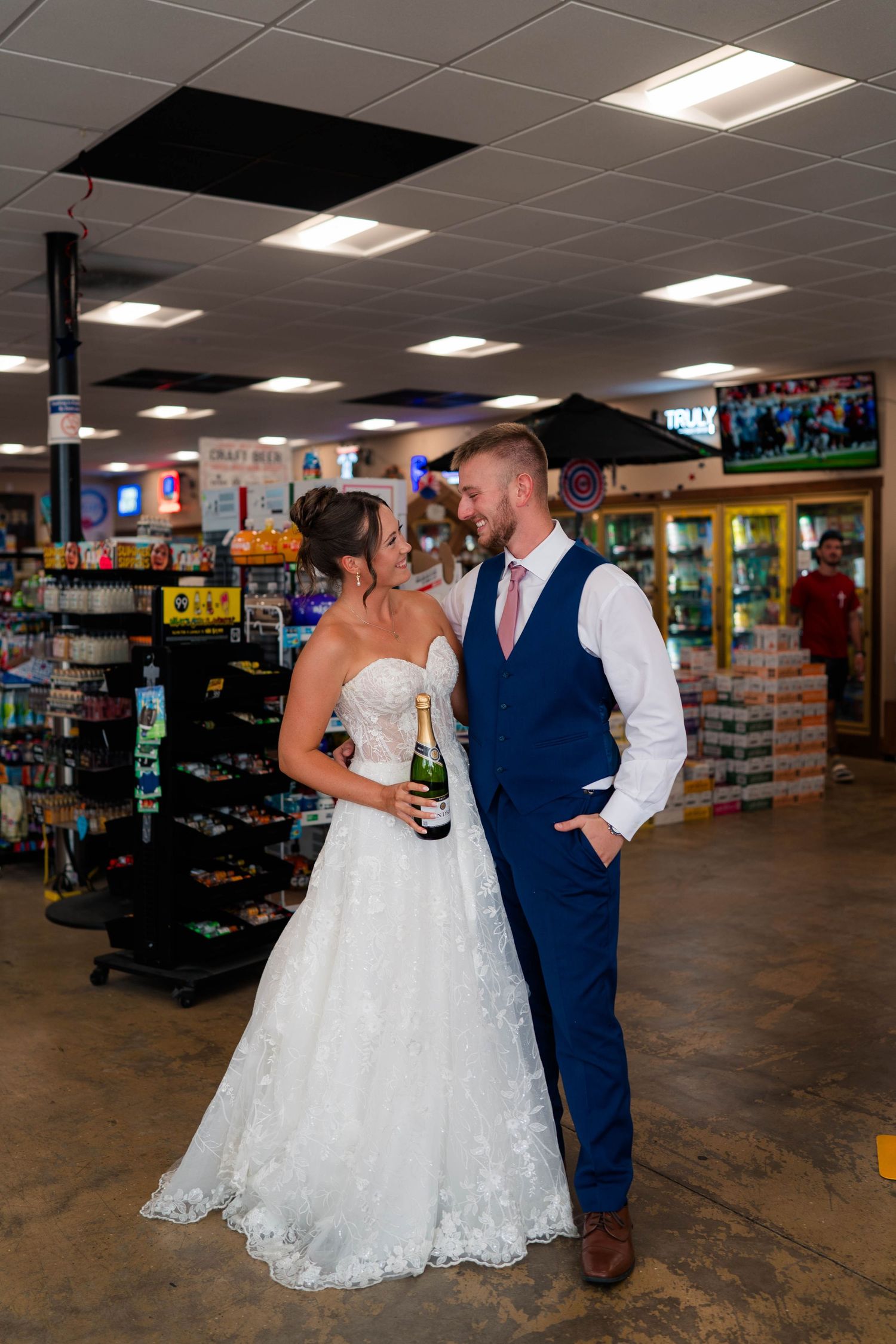 Newlyweds share a smile inside a convenience store, bride holding champagne in wedding attire.