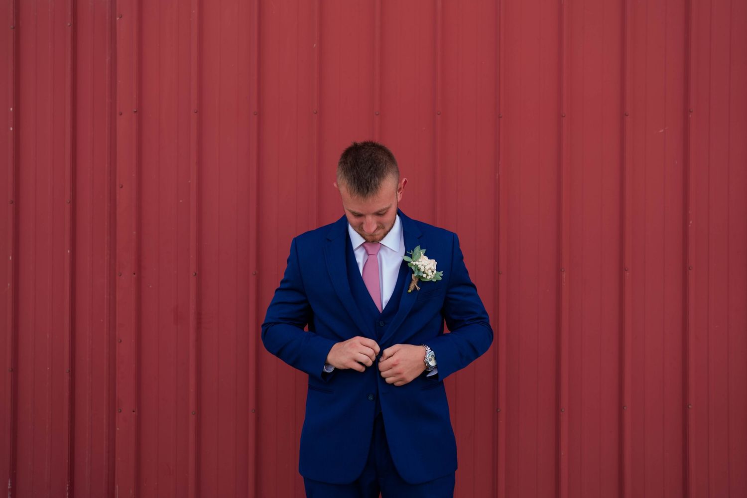 Groom in navy blue suit with pink tie and boutonniere buttoning jacket against red barn wall.