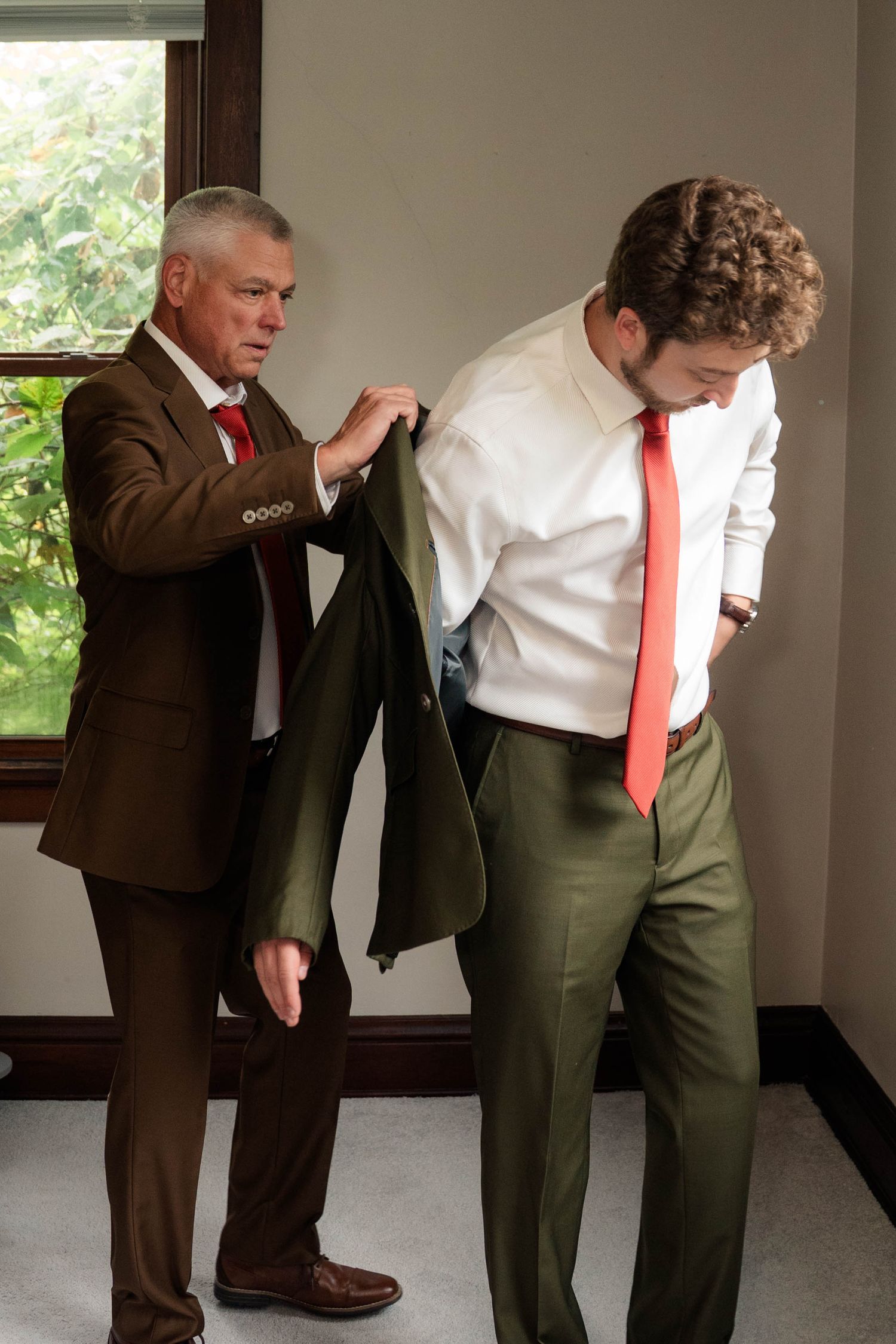 Older man in brown suit helping young groom in olive suit jacket during wedding preparations indoors.