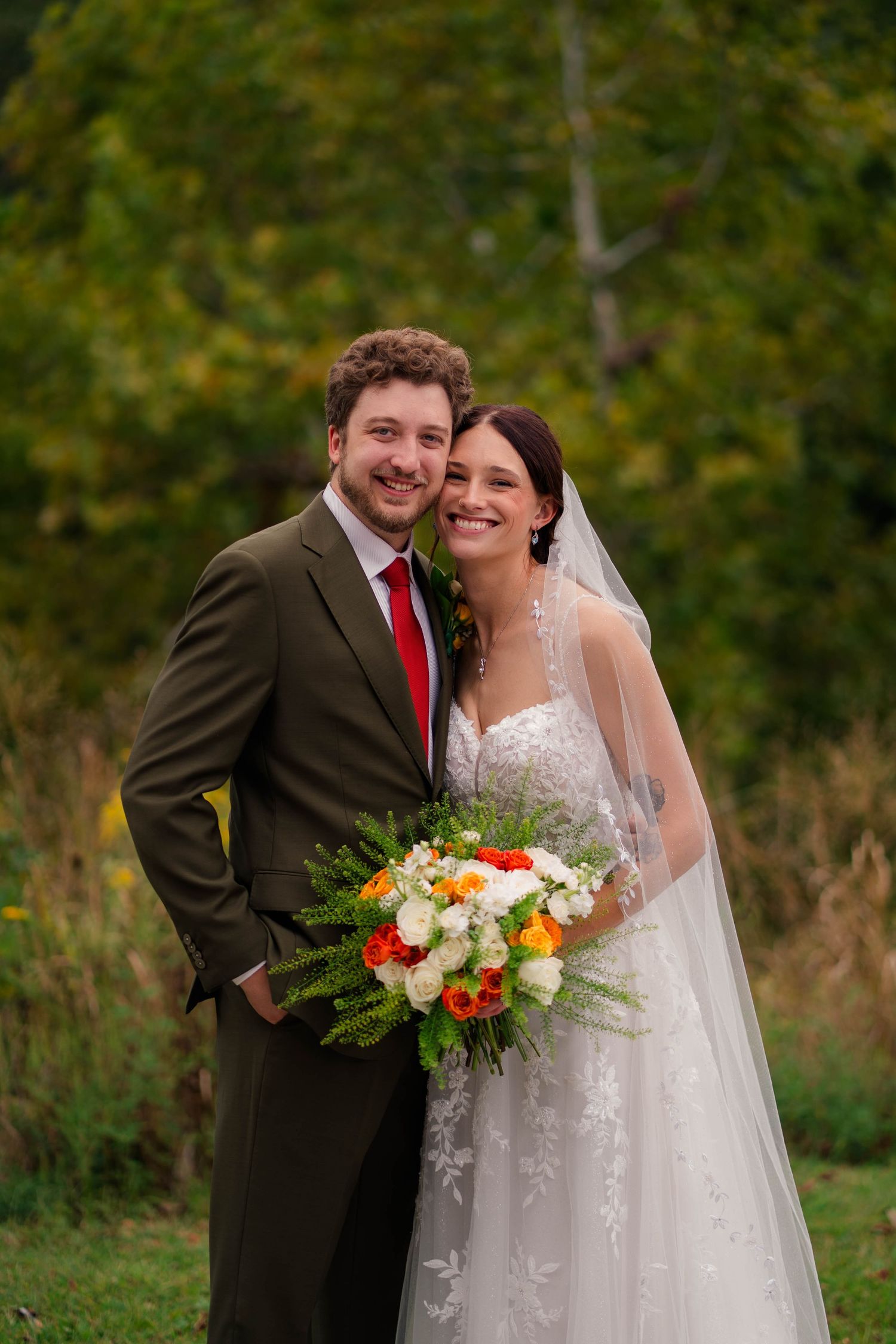Smiling bride and groom pose together outdoors holding vibrant orange and white bouquet with autumn foliage background.