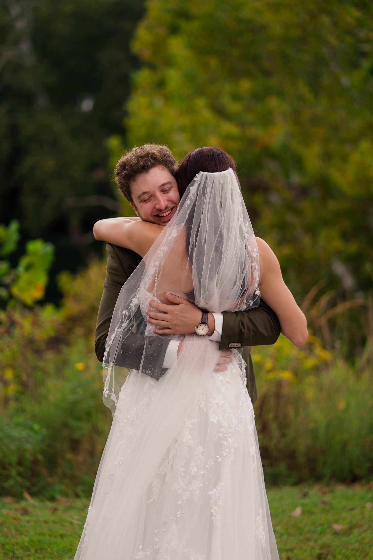Emotional groom in olive suit hugs bride in white lace gown and veil outdoors among autumn greenery.