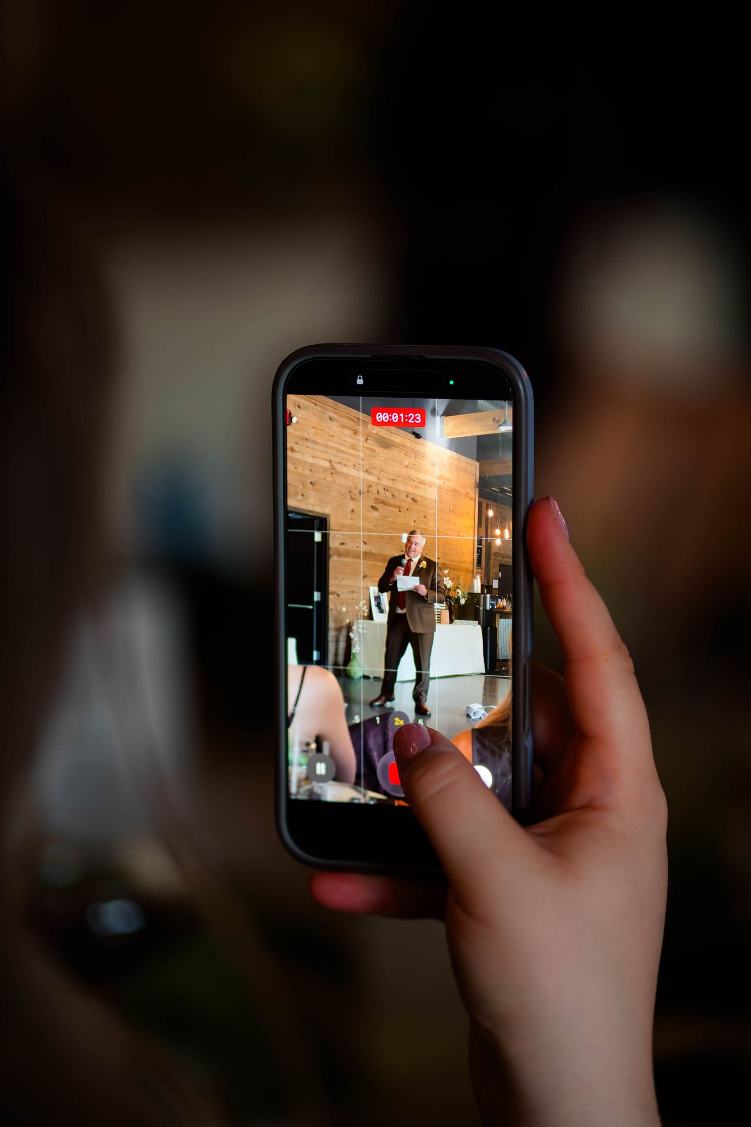 Guest recording a wedding speech on a smartphone during an indoor reception celebration.