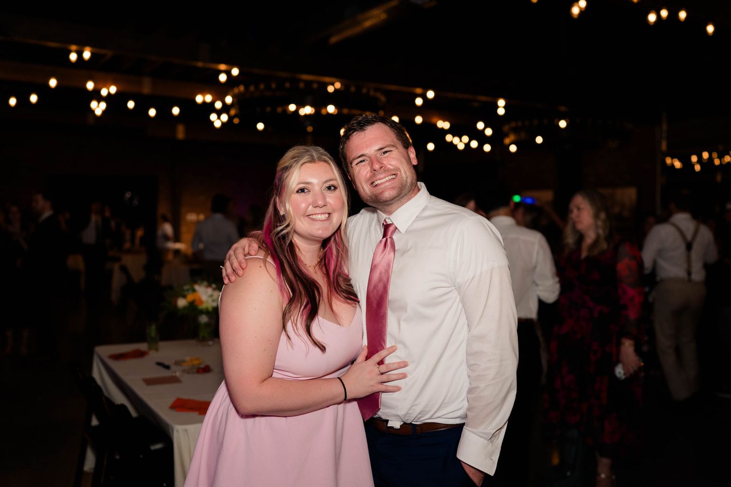 Smiling couple posing together at a wedding reception with string lights in background.