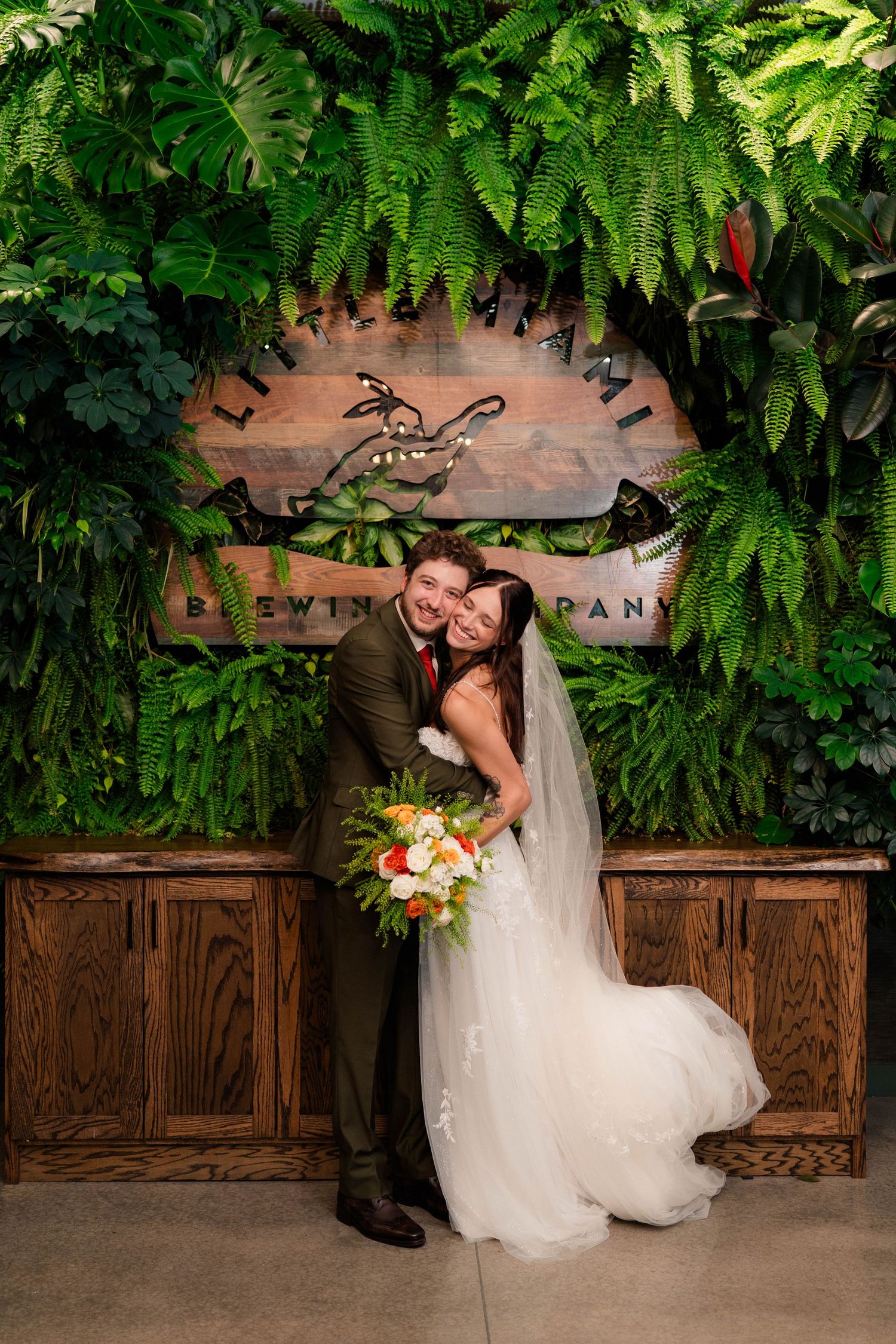 Bride and groom embrace in front of a lush green plant wall at Little Miami Brewing Company wedding.