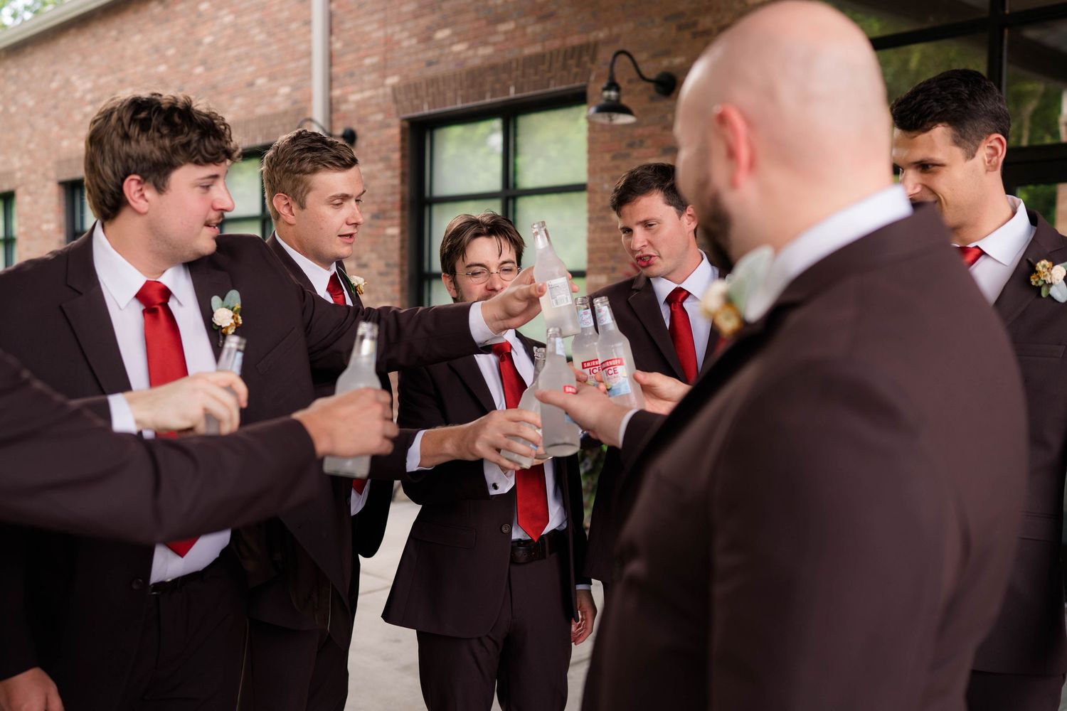 Groomsmen in dark suits with red ties toasting drinks together outside before the wedding ceremony.