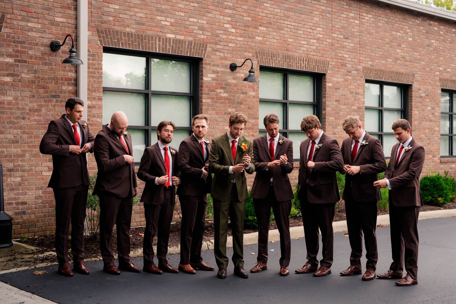 Nine groomsmen in dark suits with red ties stand in a line outside a brick building.
