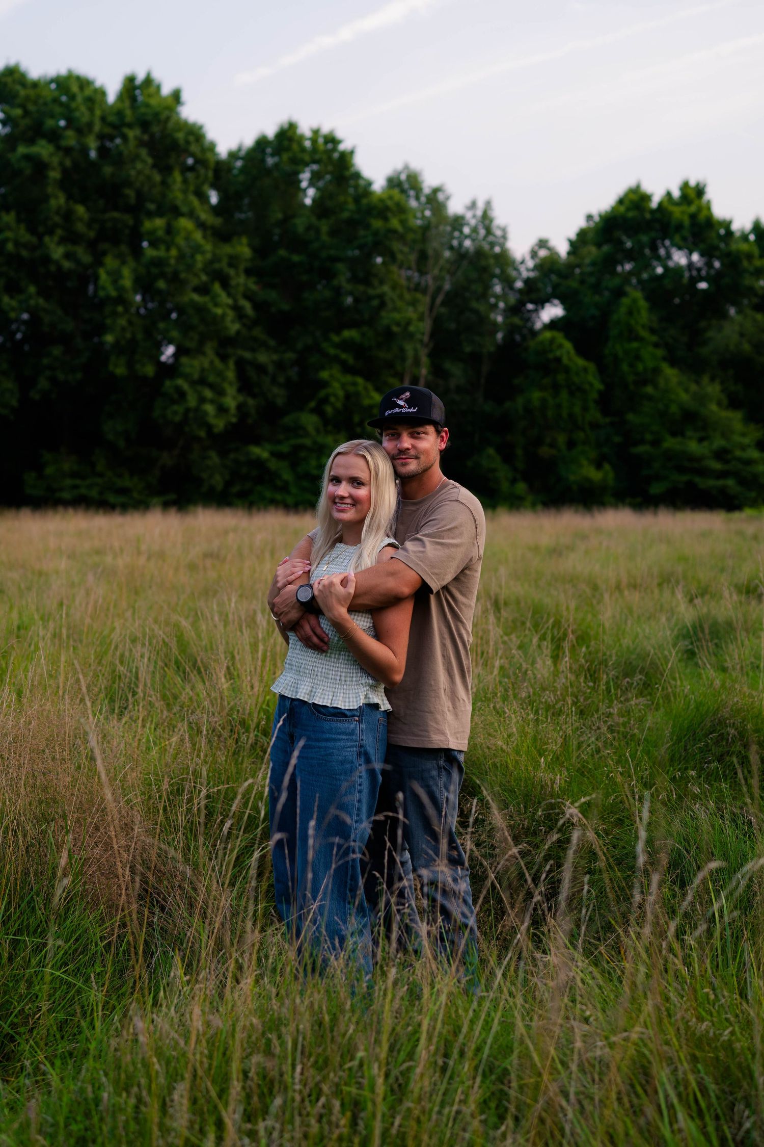 A smiling couple embraces in a tall grass meadow surrounded by green trees during golden hour.