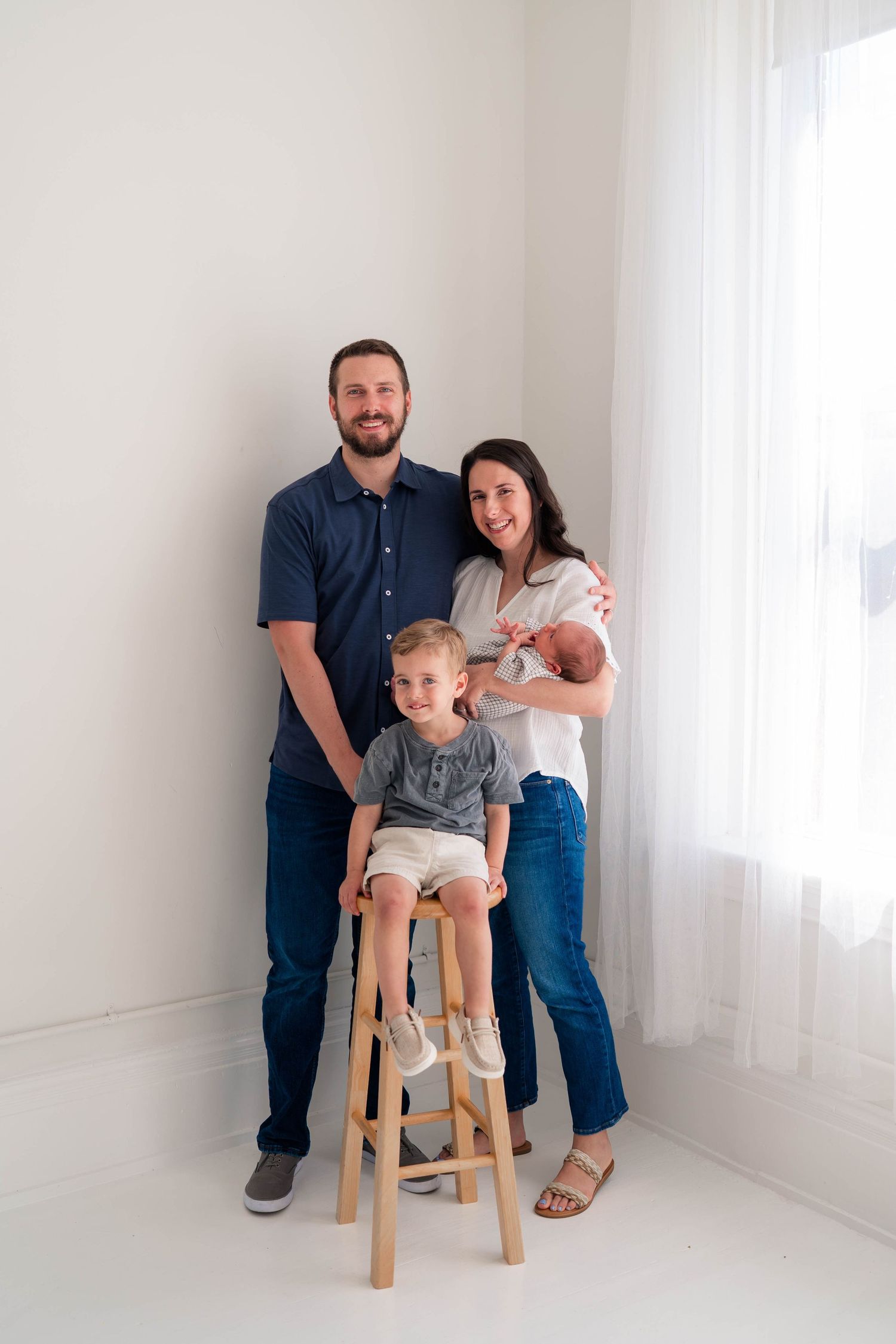 A happy family of four poses in a bright white studio, parents standing with toddler on stool and newborn in arms.