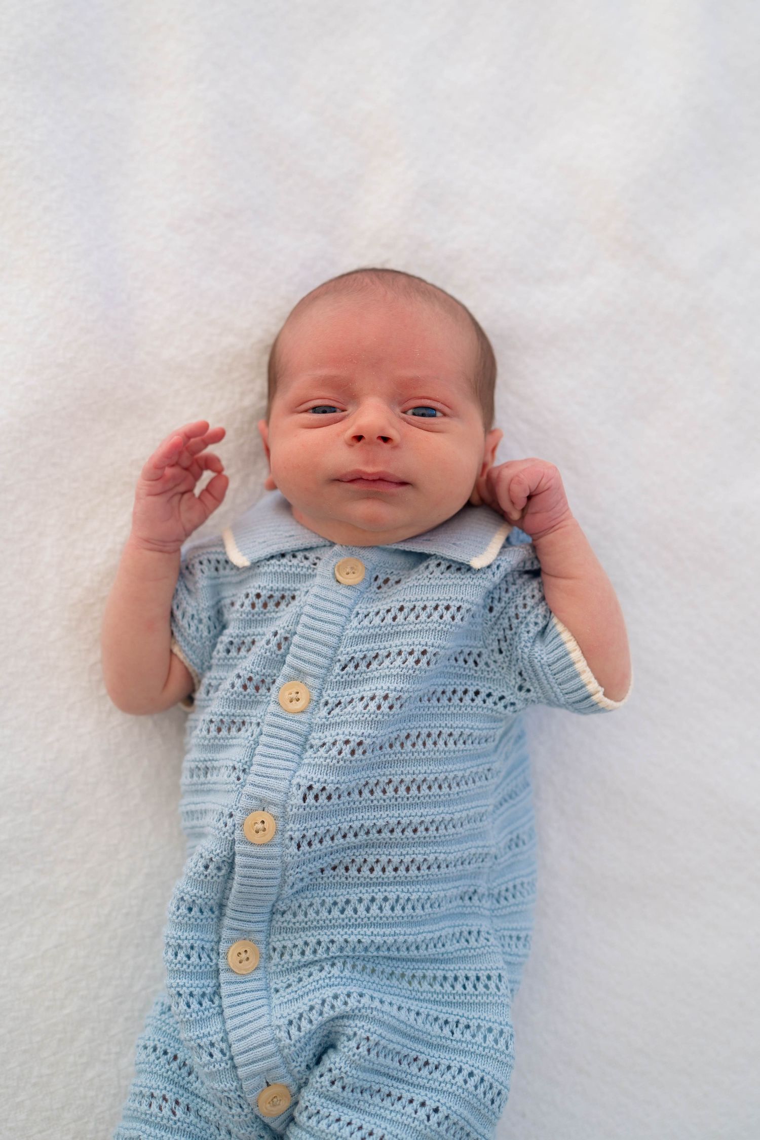 A peaceful newborn baby lies on a white blanket wearing a soft blue knit button-up romper with tiny hands raised.