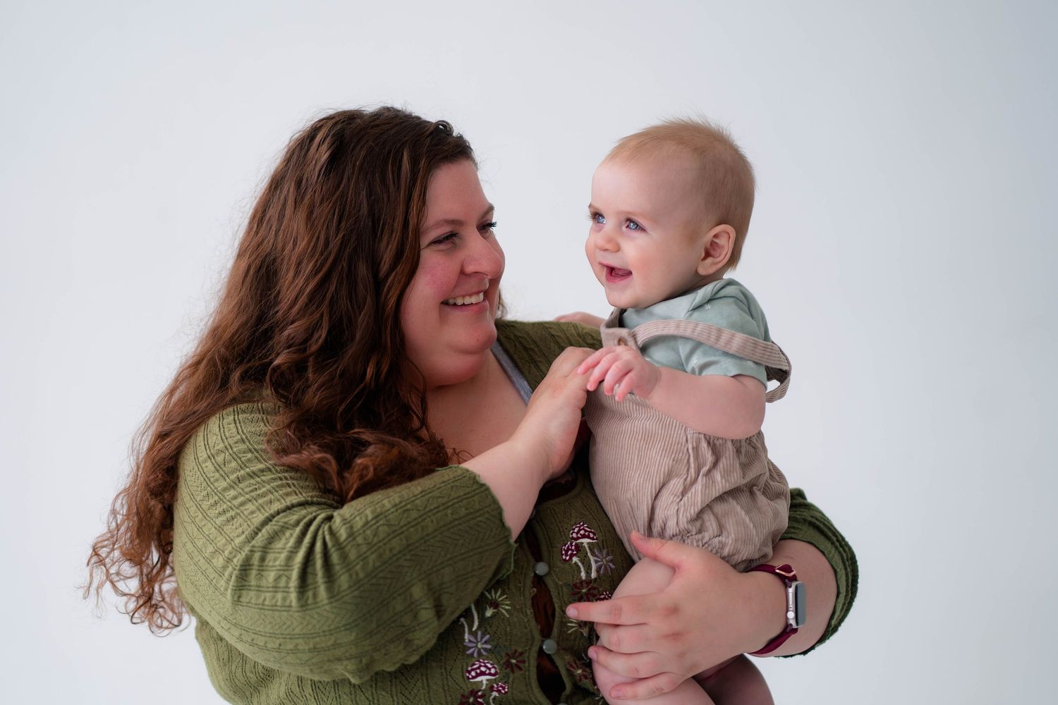 A smiling mother in a green top holds her happy laughing baby against a white studio background.