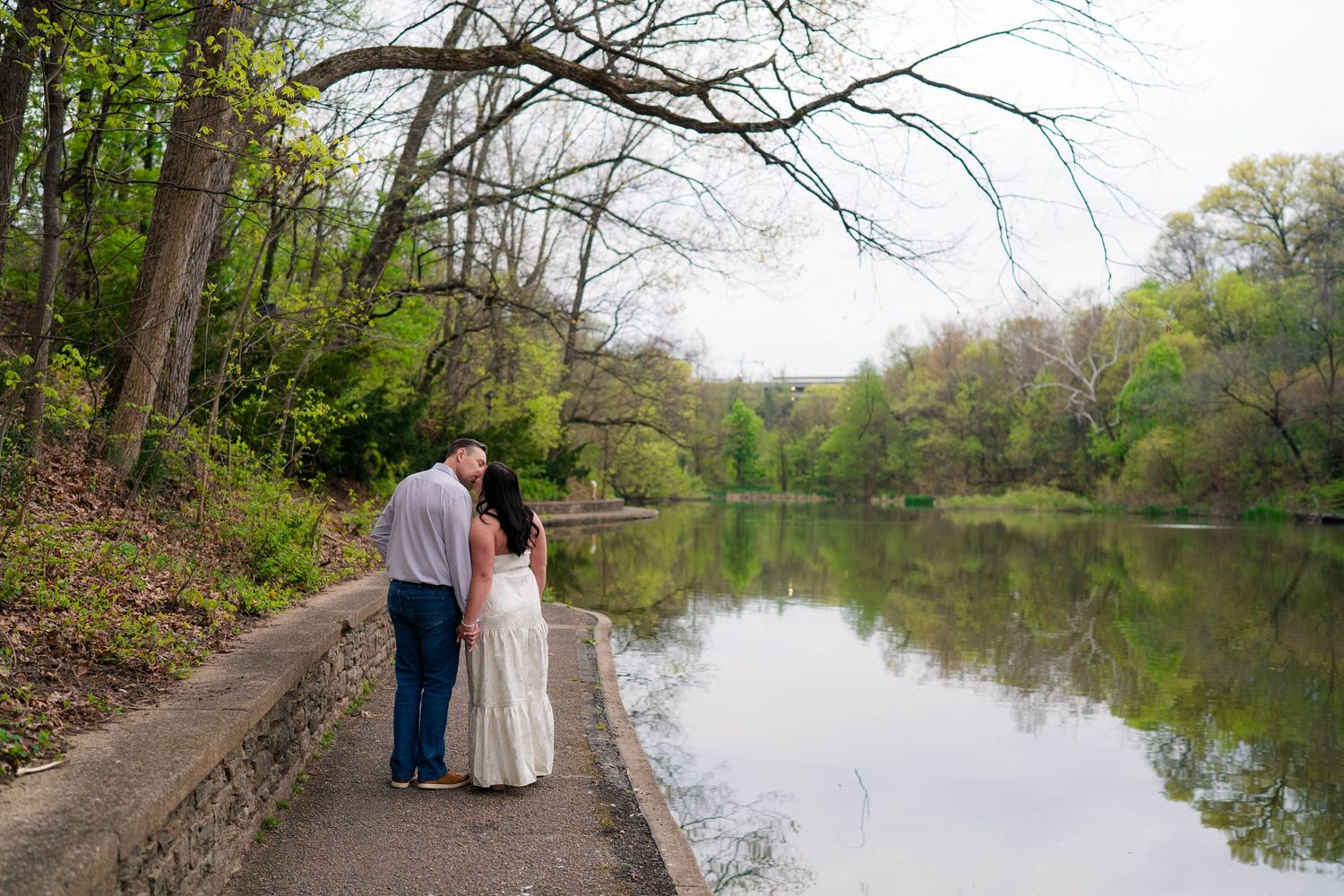 An engaged couple stands together on a lakeside path surrounded by spring trees and calm water.