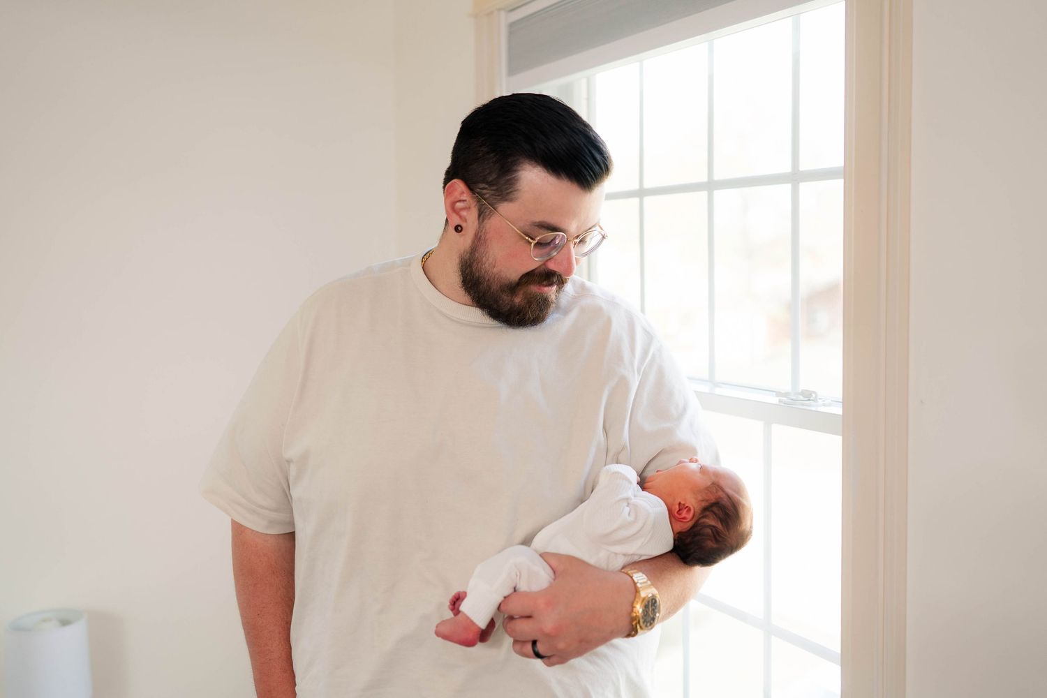 A bearded father in a white t-shirt tenderly holds his newborn baby near a bright window.