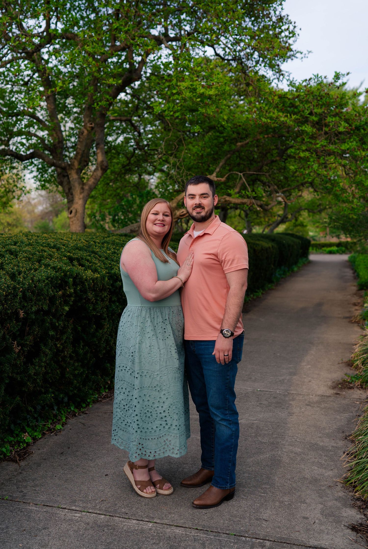 Couple posing on garden path surrounded by lush green trees and hedges, woman in mint dress, man in coral polo.