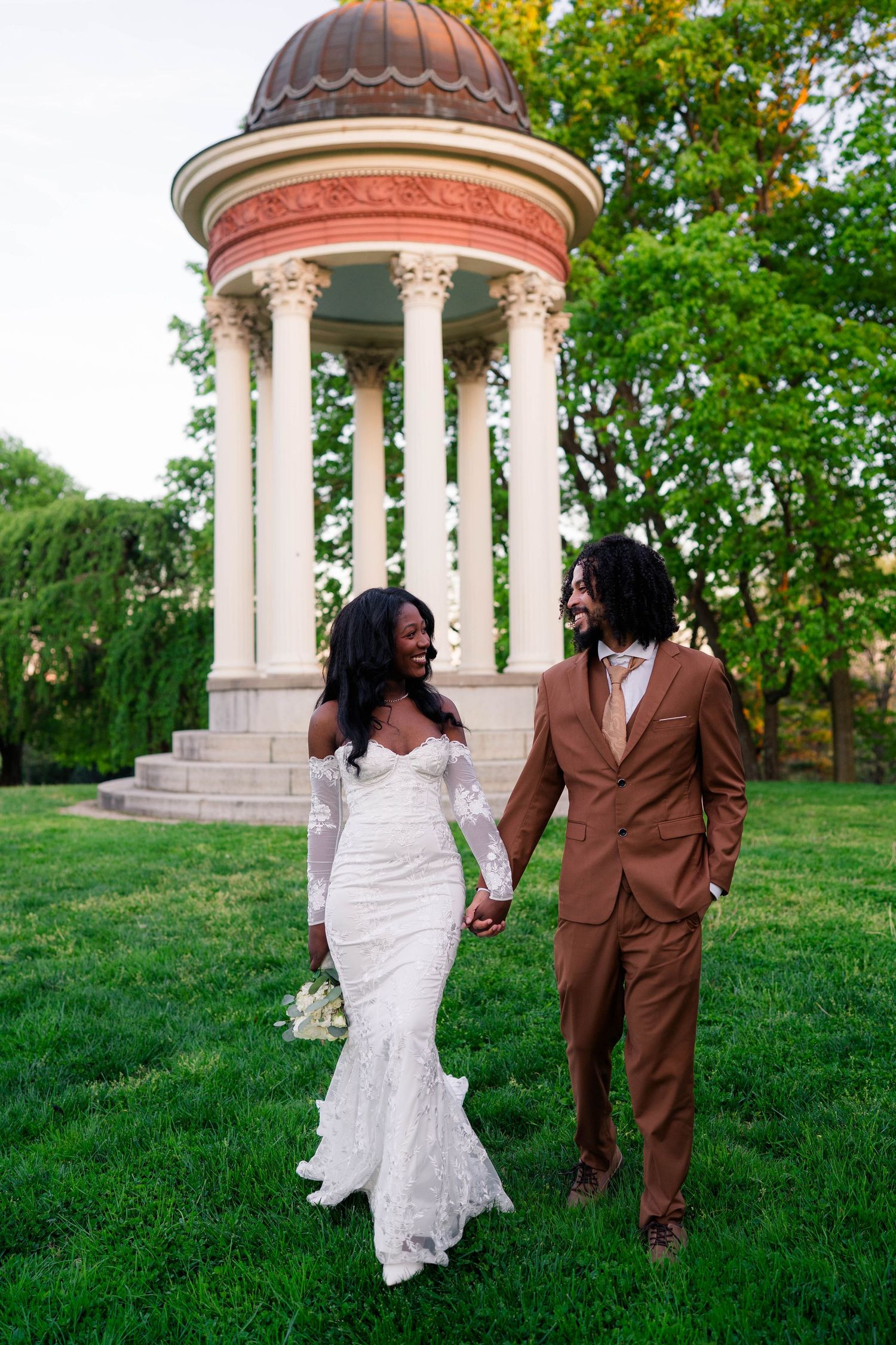 Wedding couple holds hands walking past elegant rotunda gazebo, bride in white lace gown, groom in brown suit.