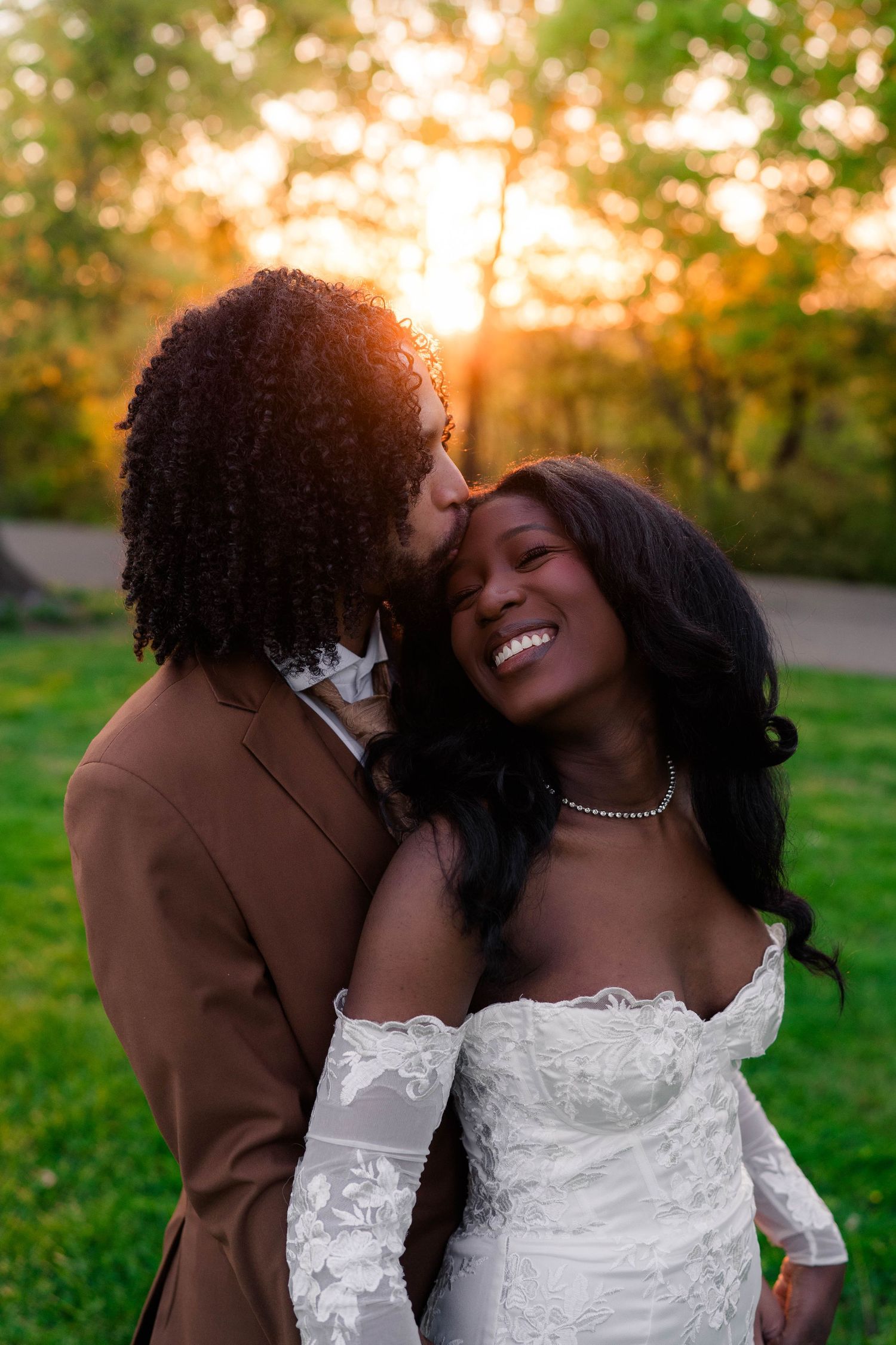 A joyful bride in a white lace gown smiles as her partner kisses her forehead during a golden sunset outdoor wedding.