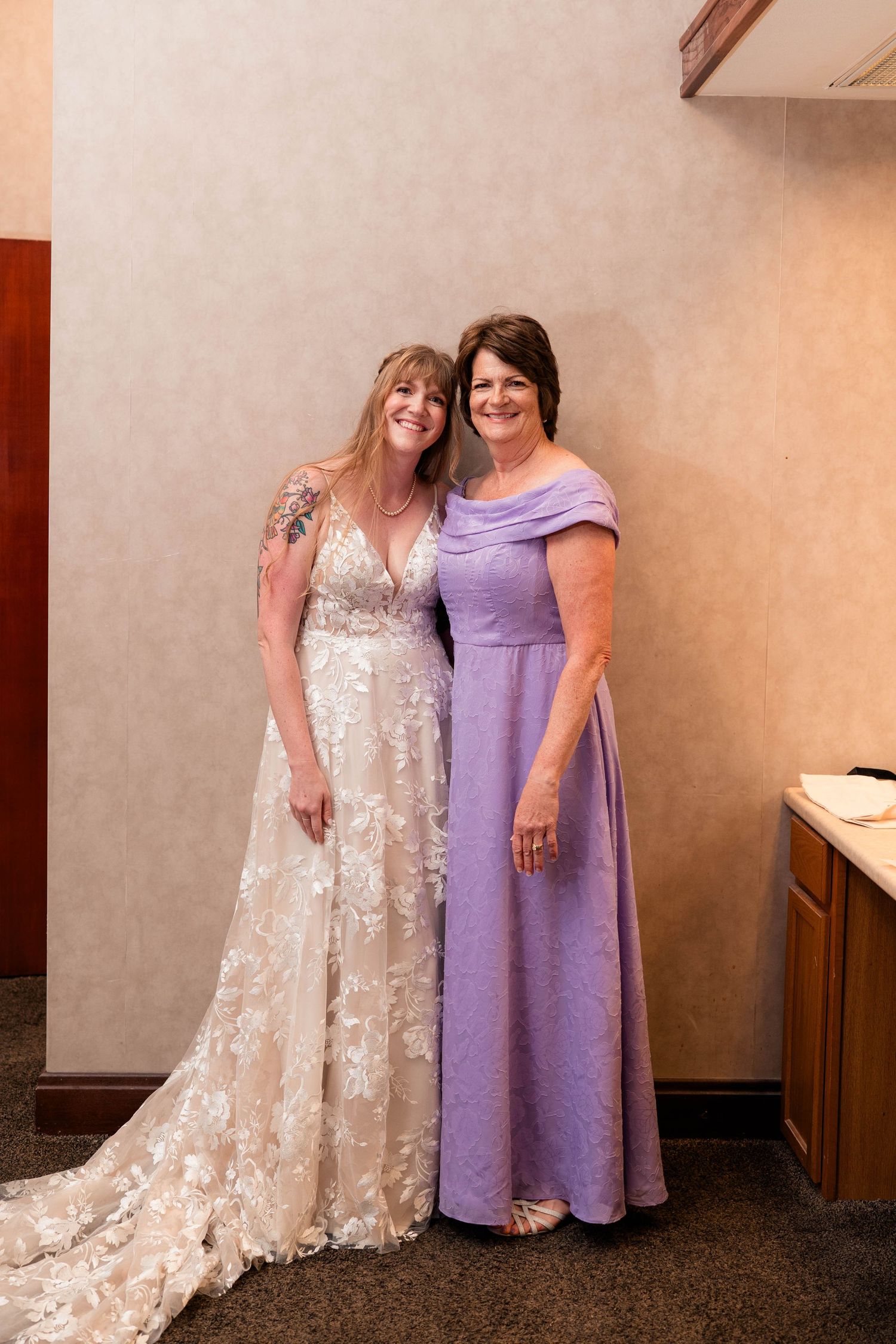 Bride in lace wedding gown poses with woman in lavender off-shoulder gown against neutral wall.