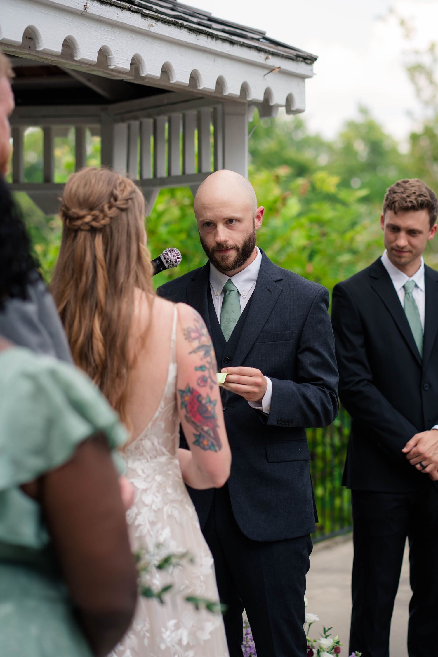 Bald bearded groom in dark suit reads vows while holding a small card during outdoor gazebo wedding ceremony.