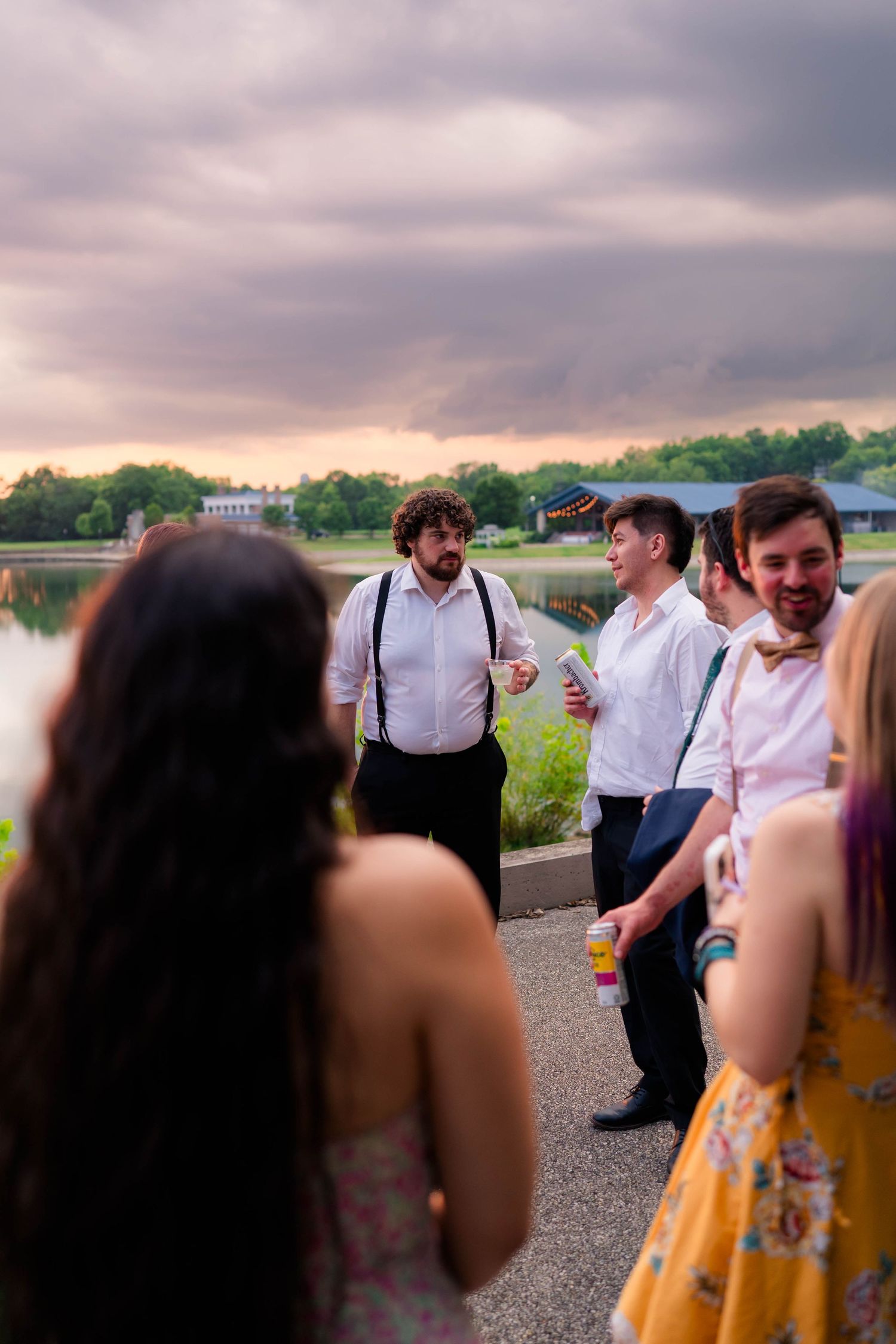 Wedding guests socializing outdoors near lake under dramatic cloudy sunset sky.