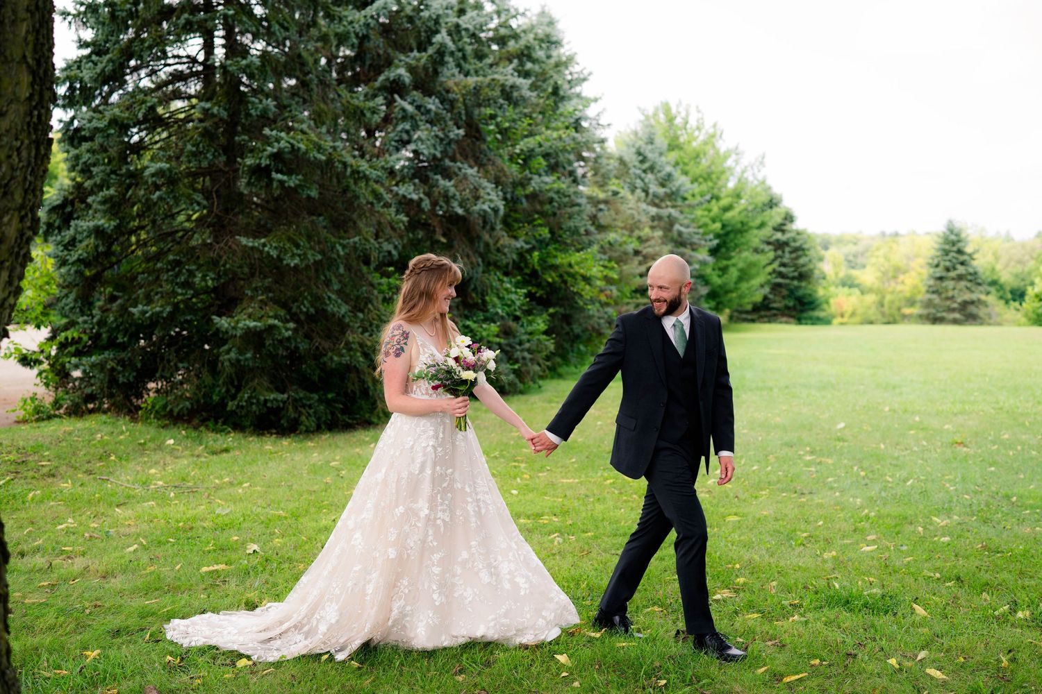 Bride and groom walk hand in hand across green lawn with tall trees.