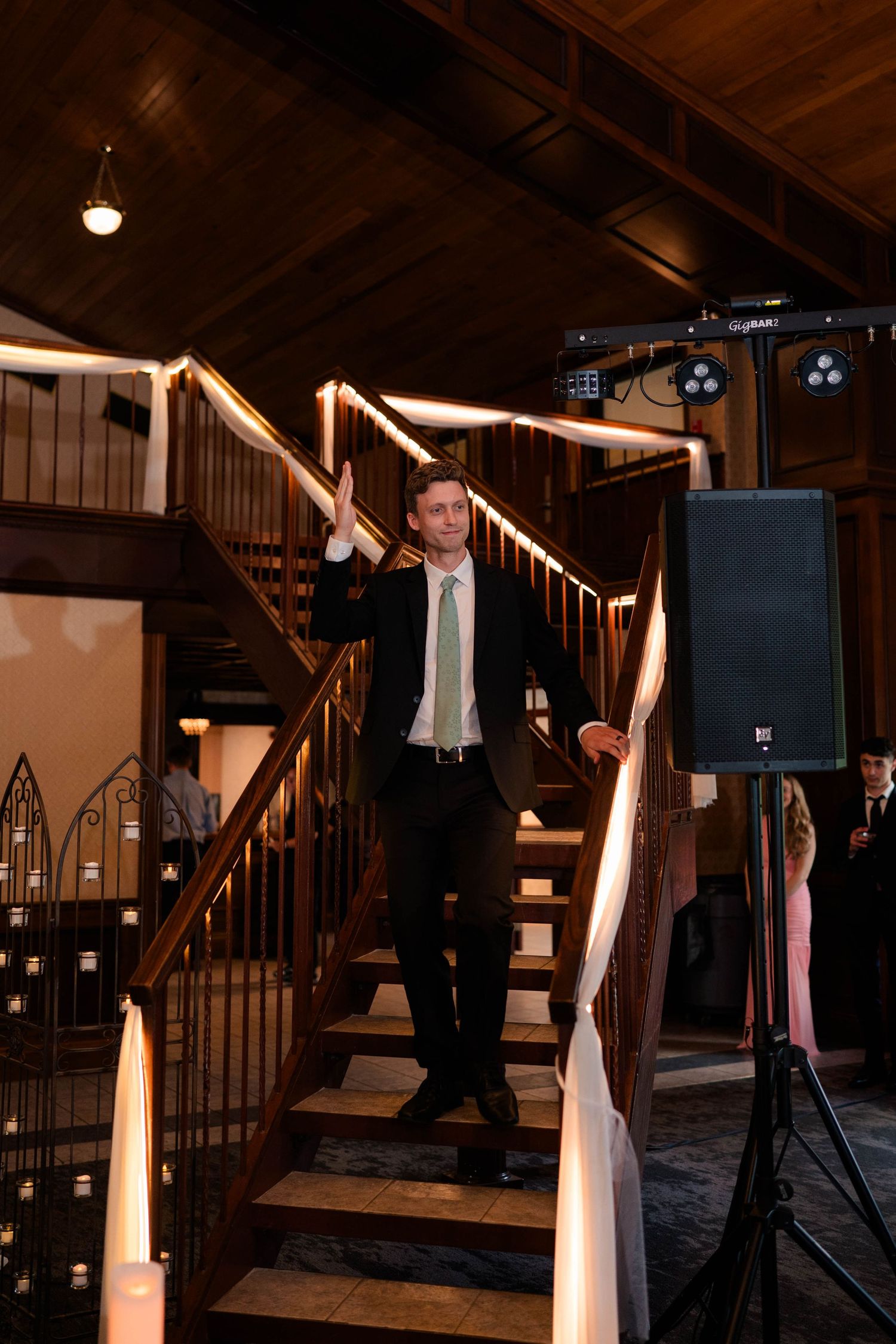 Groomsman in dark suit and green tie waves while descending decorated staircase at indoor wedding reception venue.