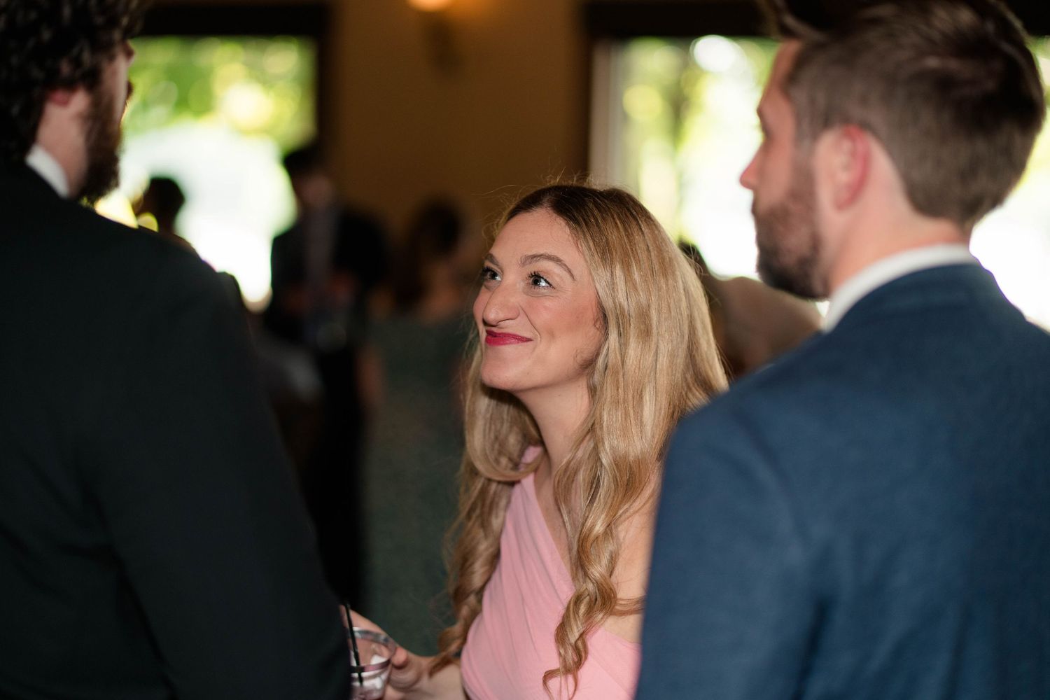 Blonde woman in pink dress looks up smiling while conversing at indoor wedding reception.