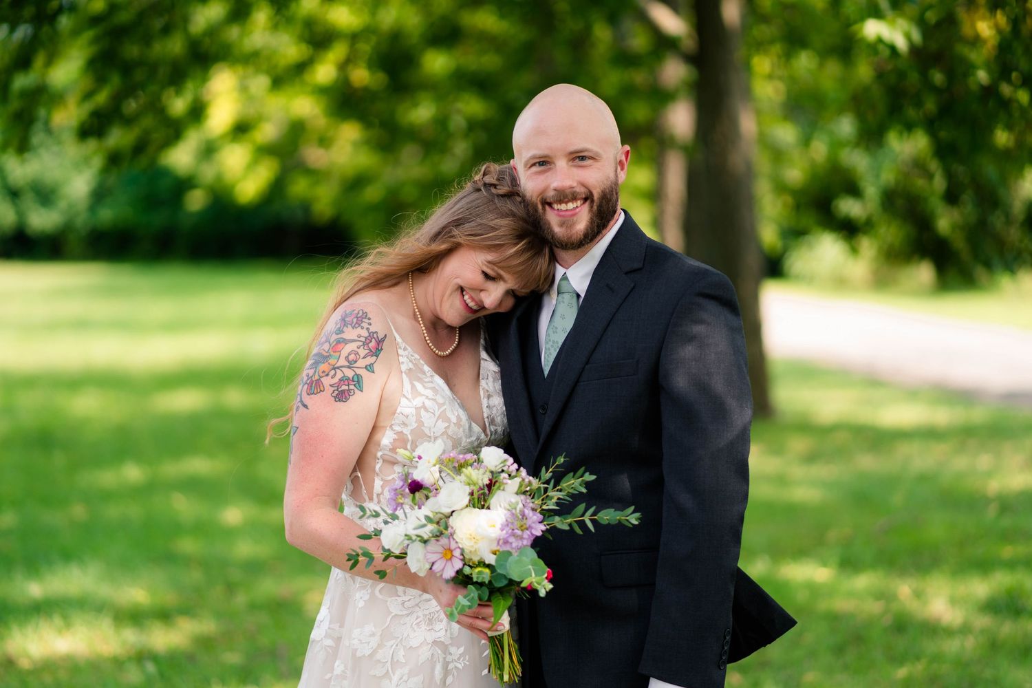 Smiling bald groom embraces tattooed bride holding white and purple bouquet in sunny green park.