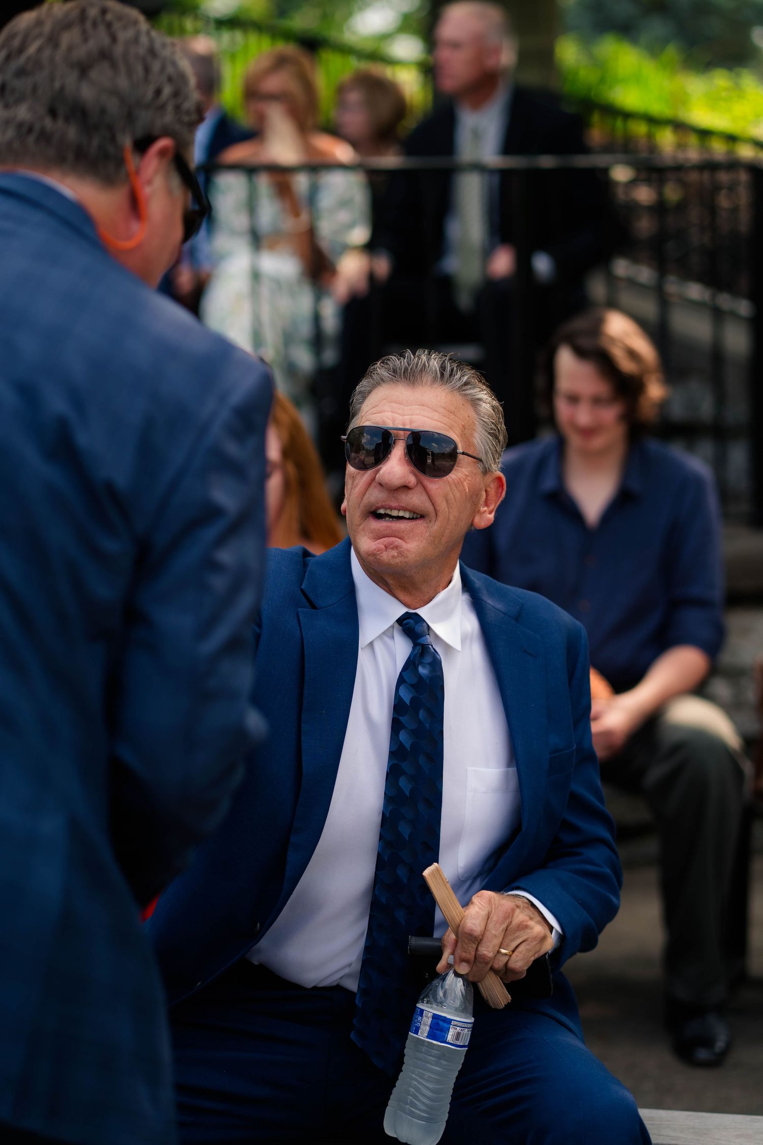 Older man in blue suit and sunglasses smiles while chatting at outdoor wedding reception.