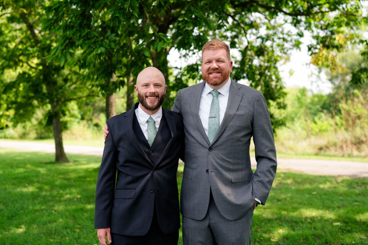 Two smiling groomsmen in dark suits pose together outdoors under green trees.