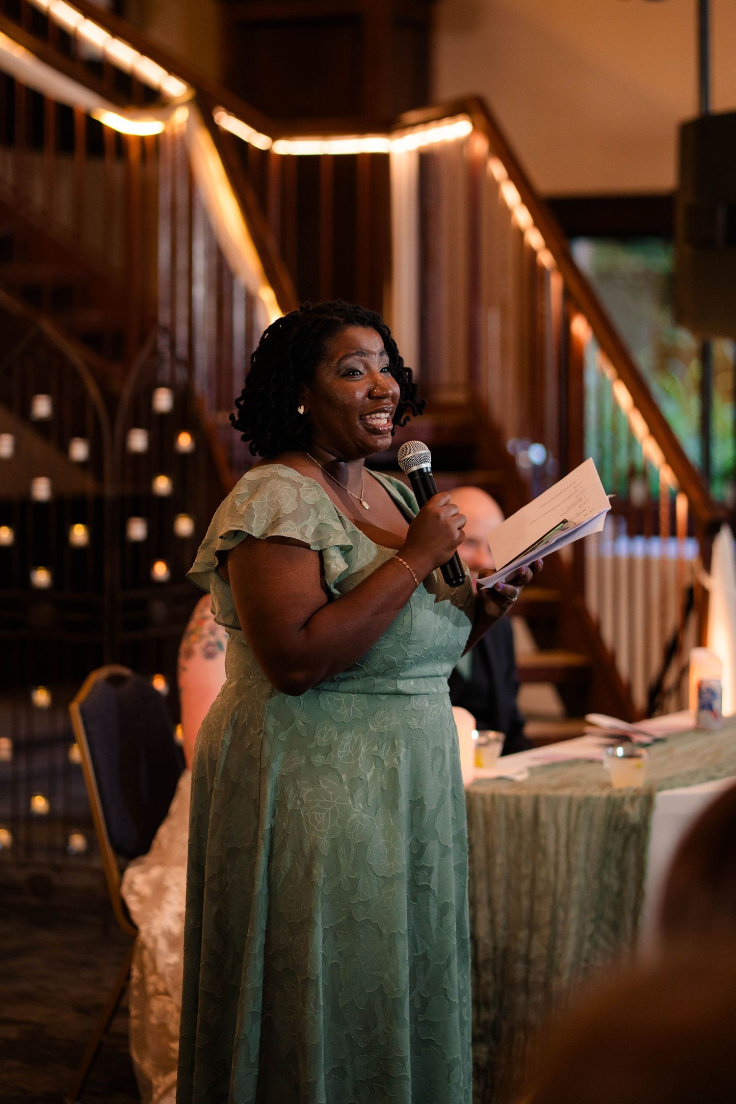 Woman in sage green dress smiles while giving speech with microphone at elegant wedding reception.