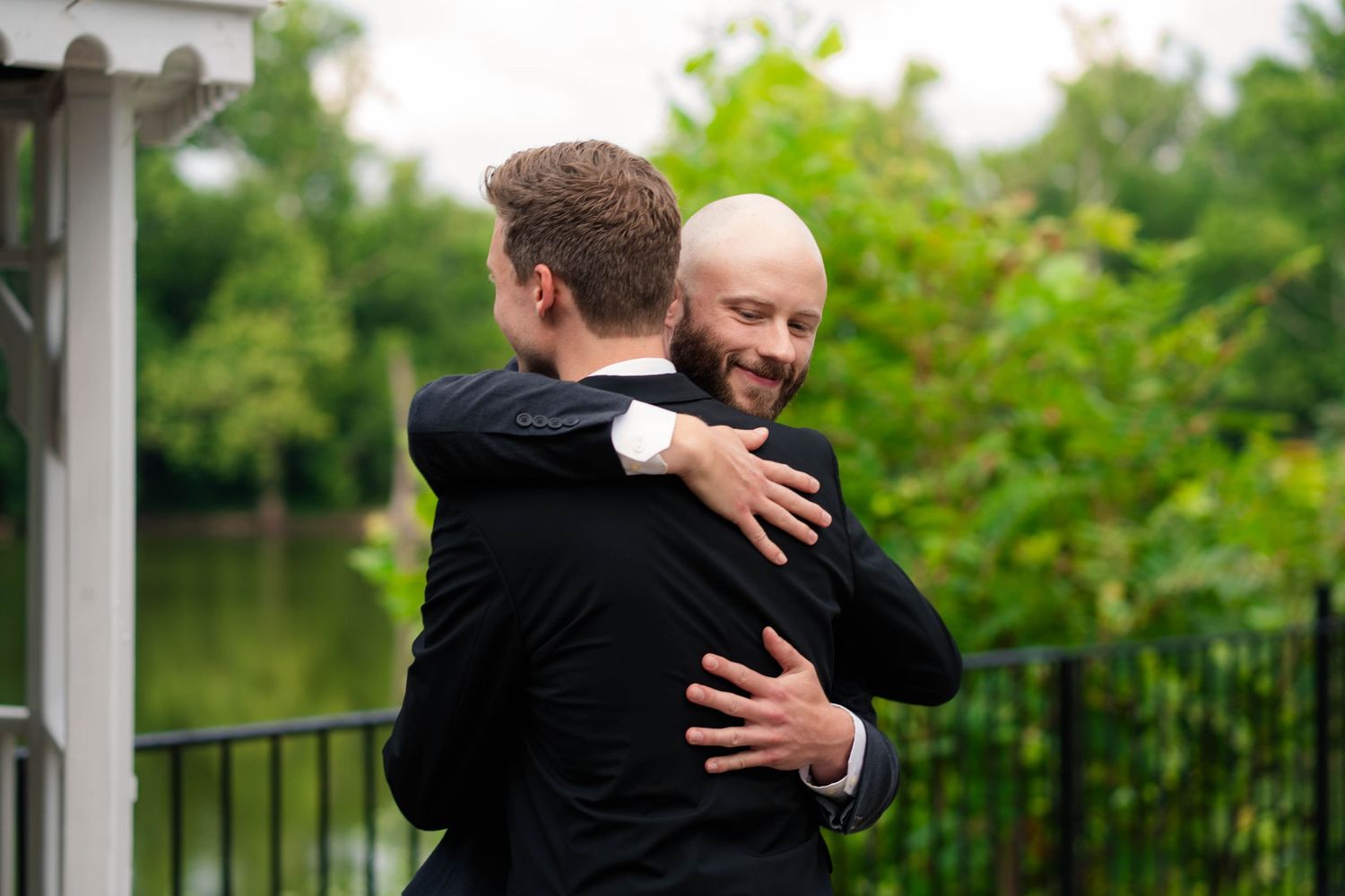 Bald groom smiles while embracing groomsman in black suit near scenic waterfront backdrop.