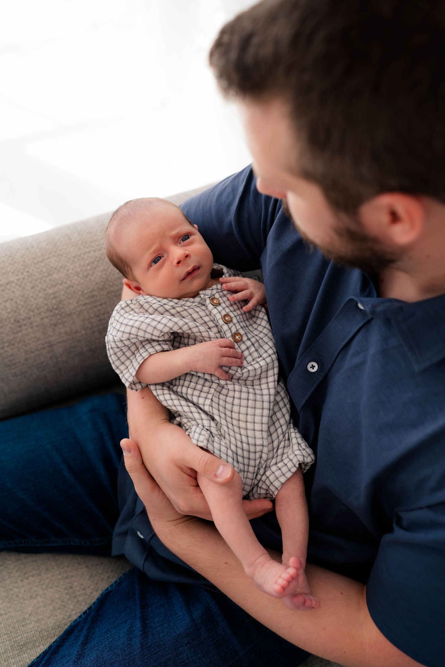 Proud father in navy shirt cradling his alert newborn baby dressed in a plaid romper on a gray sofa.