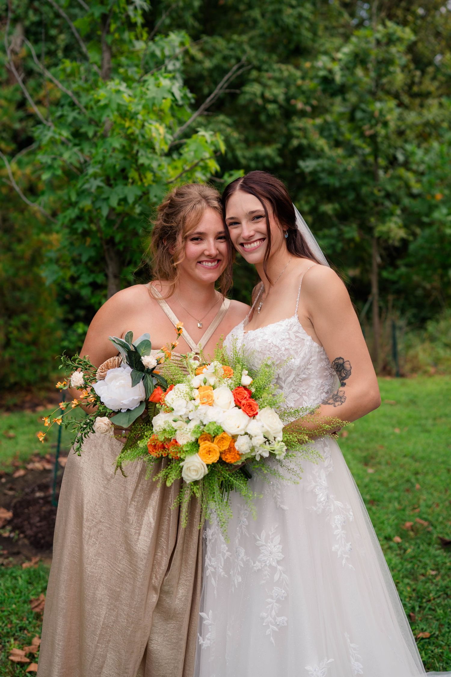 Smiling bride and bridesmaid pose together outdoors holding colorful orange and white wedding bouquets.