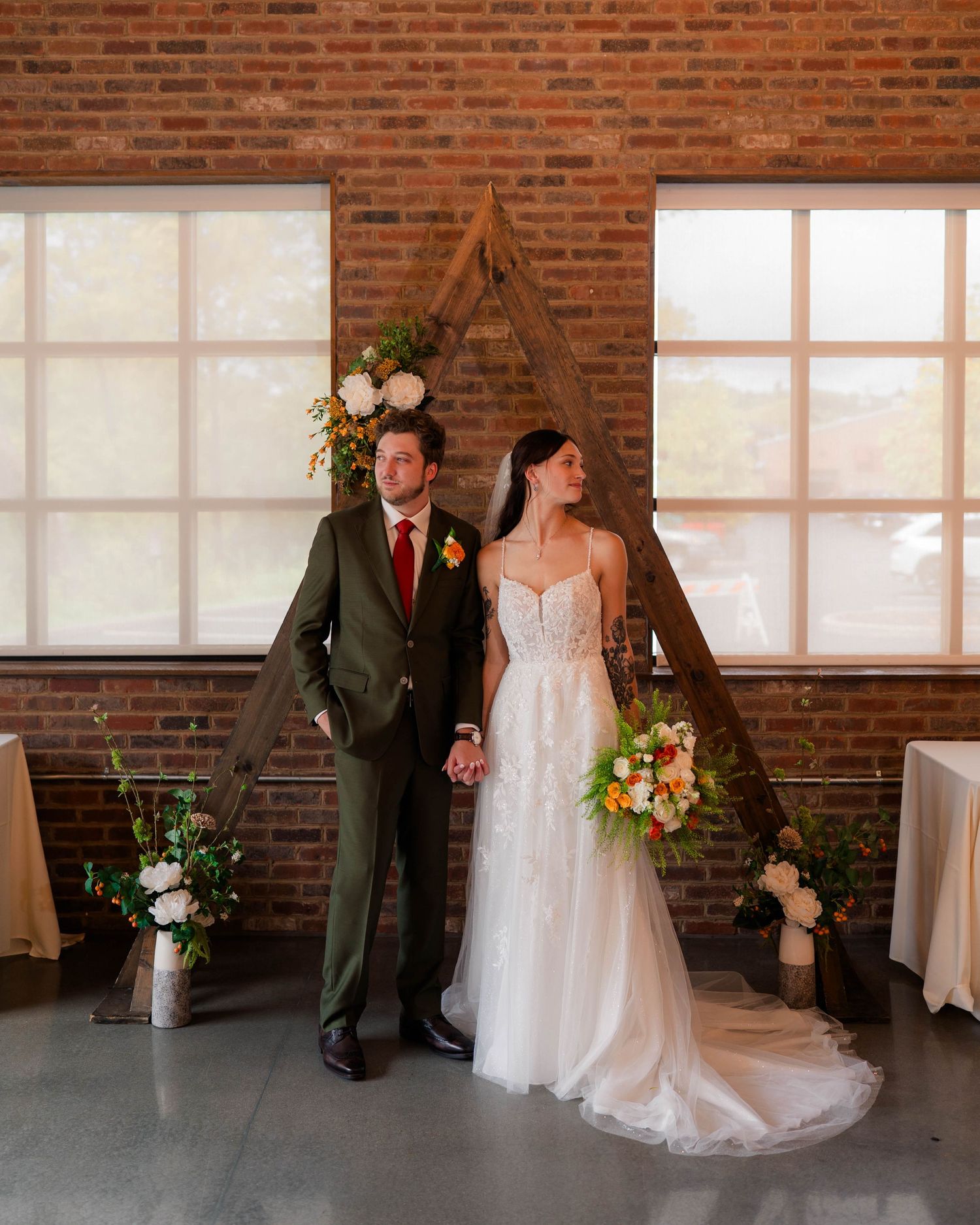 Bride and groom posing before wooden triangle arch with floral decor in brick venue.