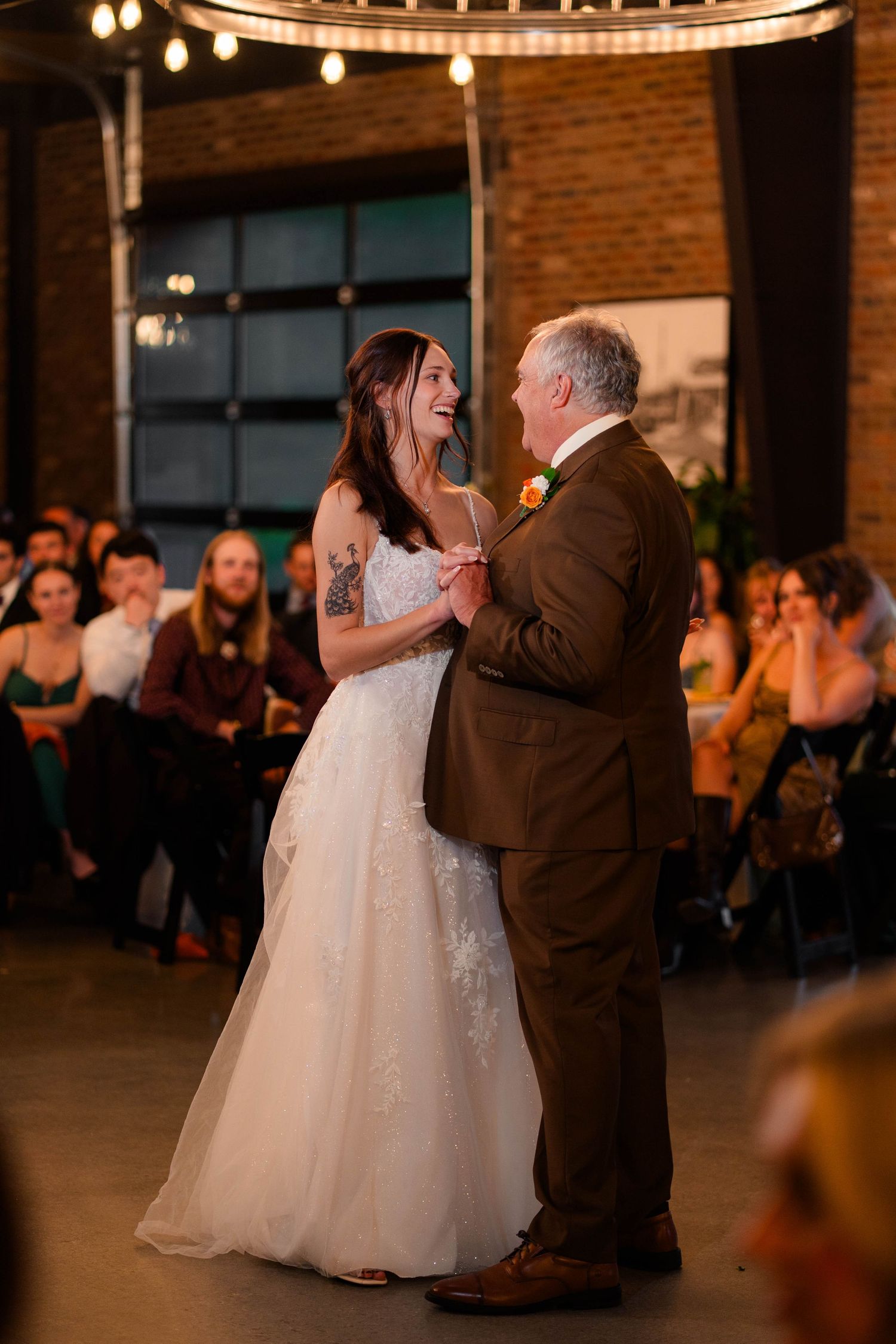 Bride in white gown shares joyful father-daughter dance under warm lights at rustic industrial wedding reception.