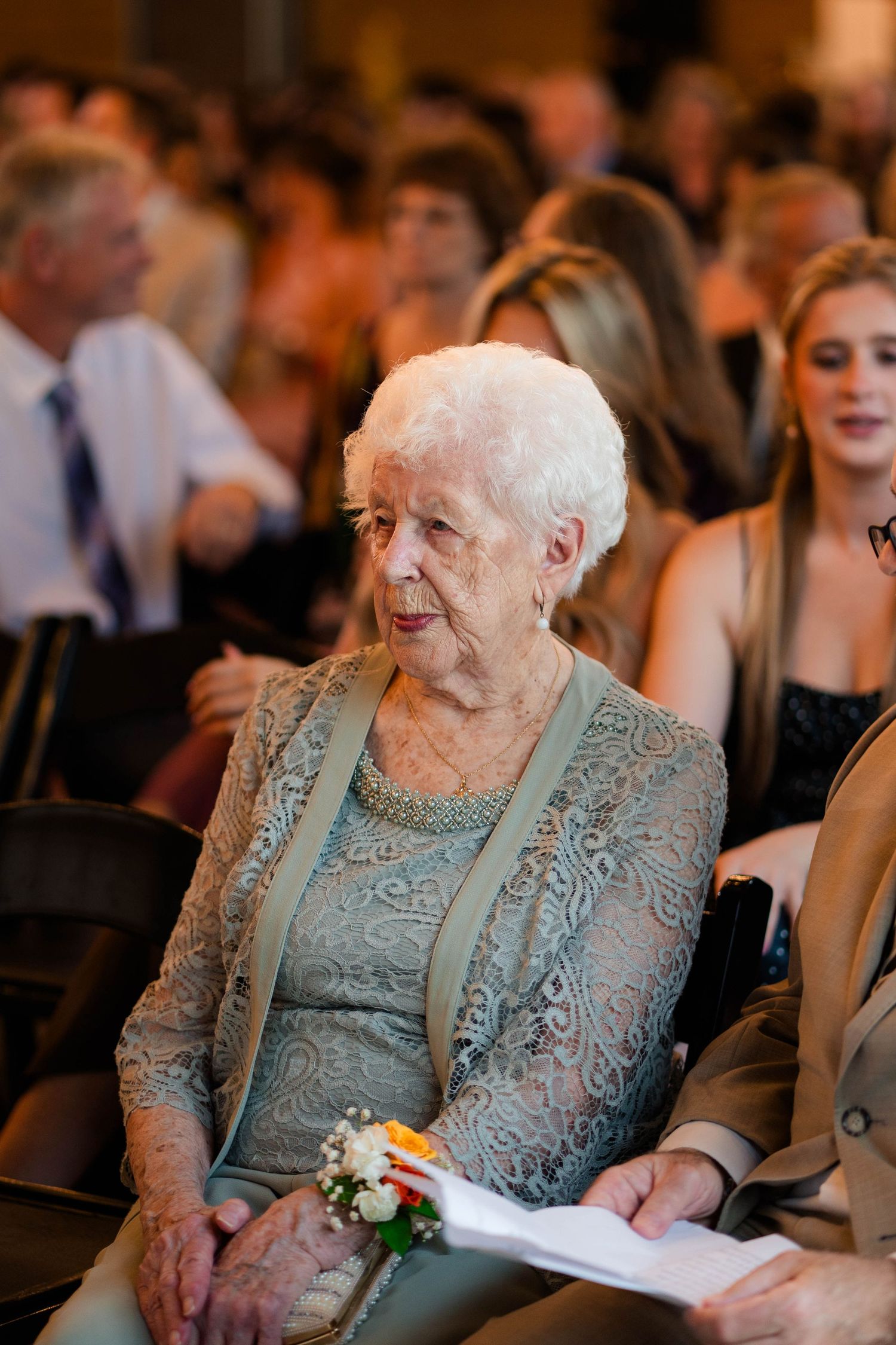 Elderly wedding guest in sage lace dress sits attentively holding program at wedding ceremony.