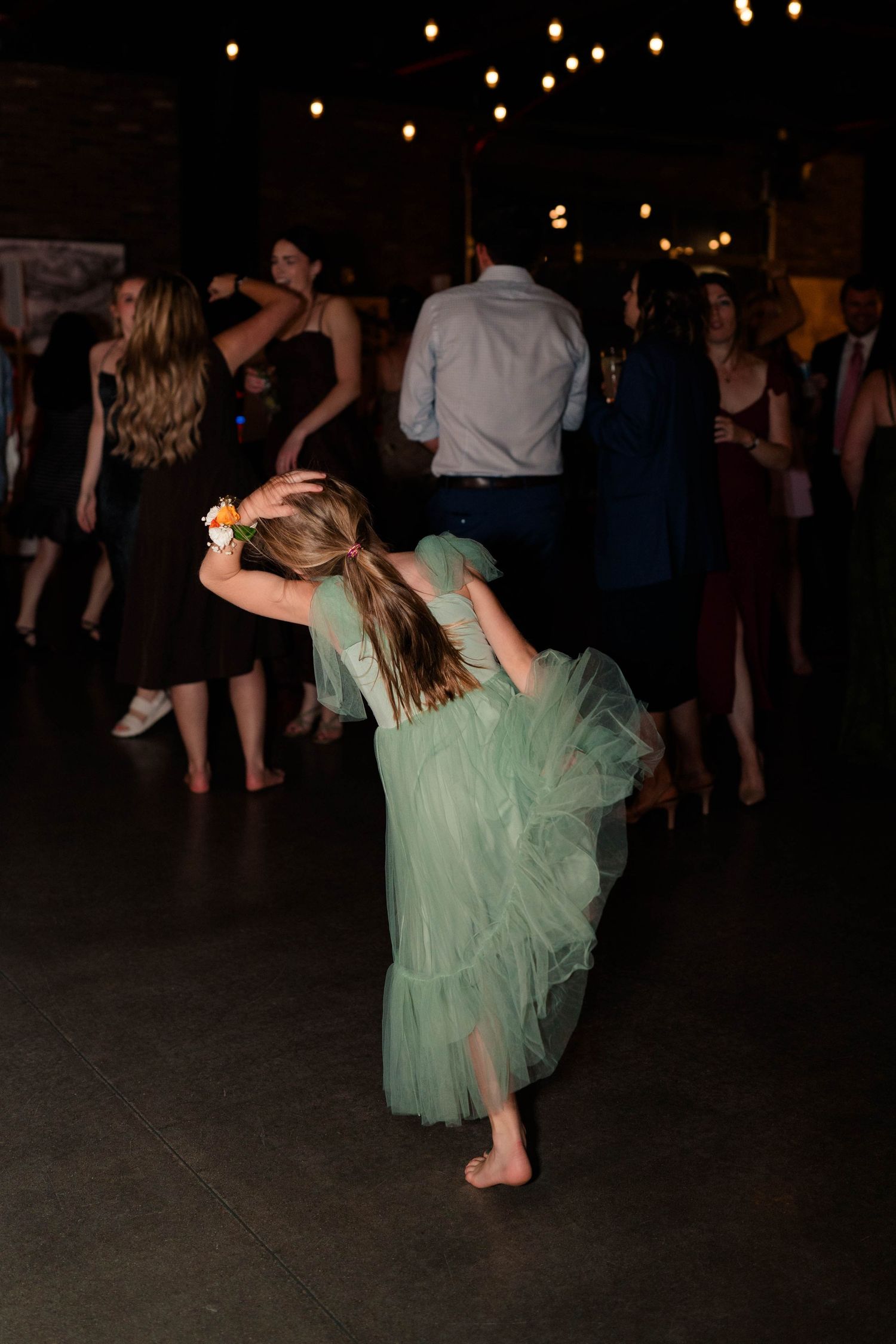 Young girl in a mint green tulle dress dancing barefoot at a wedding reception under string lights.