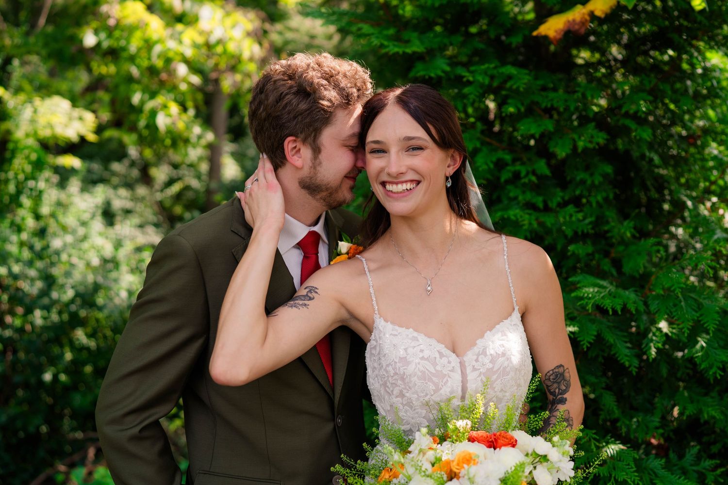 Groom whispers to smiling bride holding colorful bouquet, surrounded by green foliage outdoors.