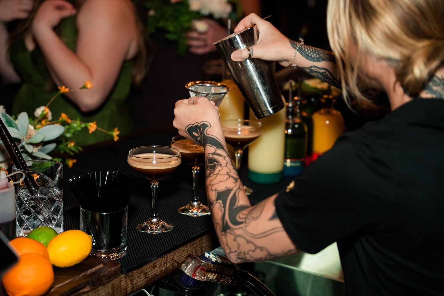 Tattooed bartender pouring espresso martinis at wedding reception bar with citrus garnishes.