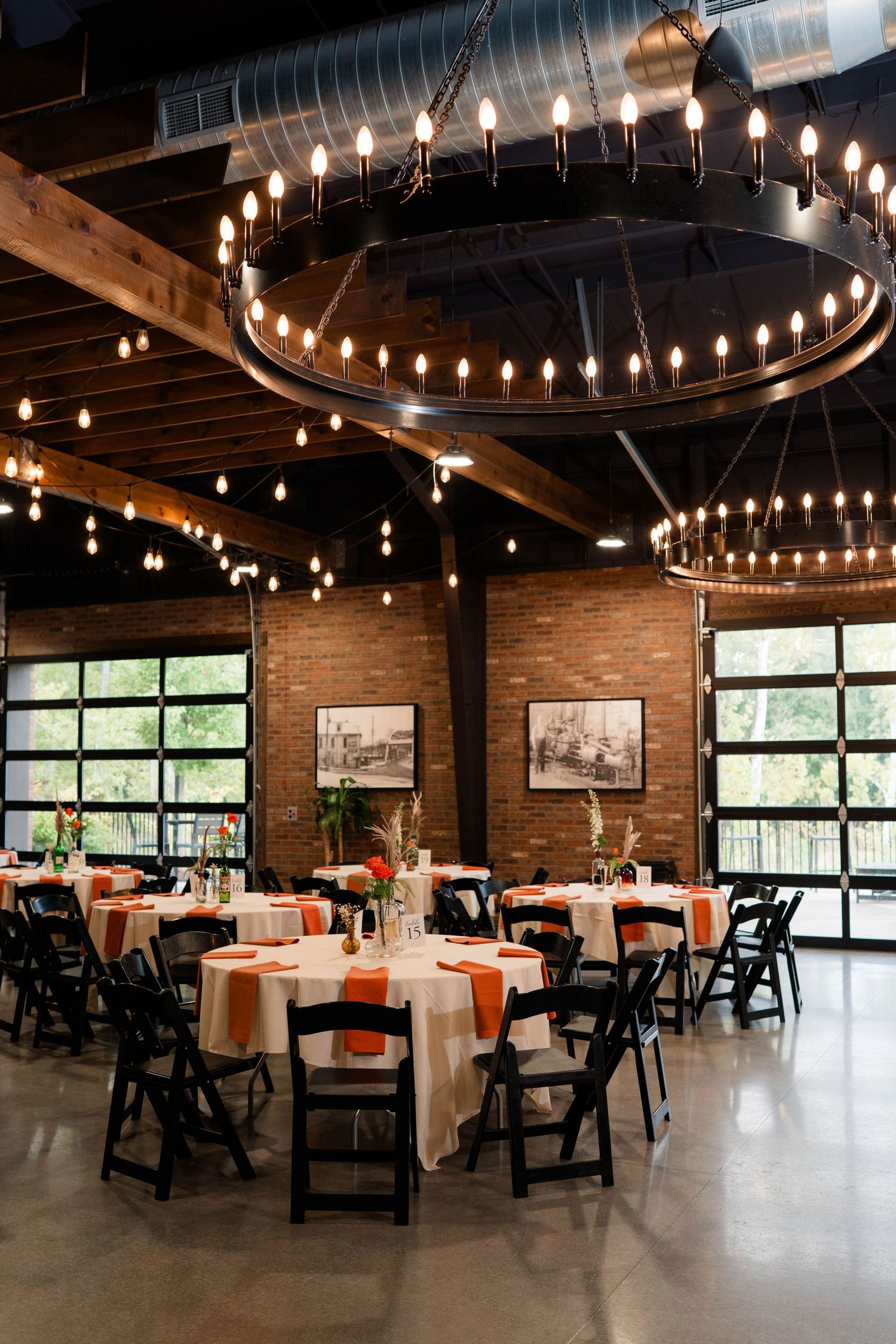 Rustic wedding reception space with barrel chandelier, string lights, orange table linens, and exposed brick walls.