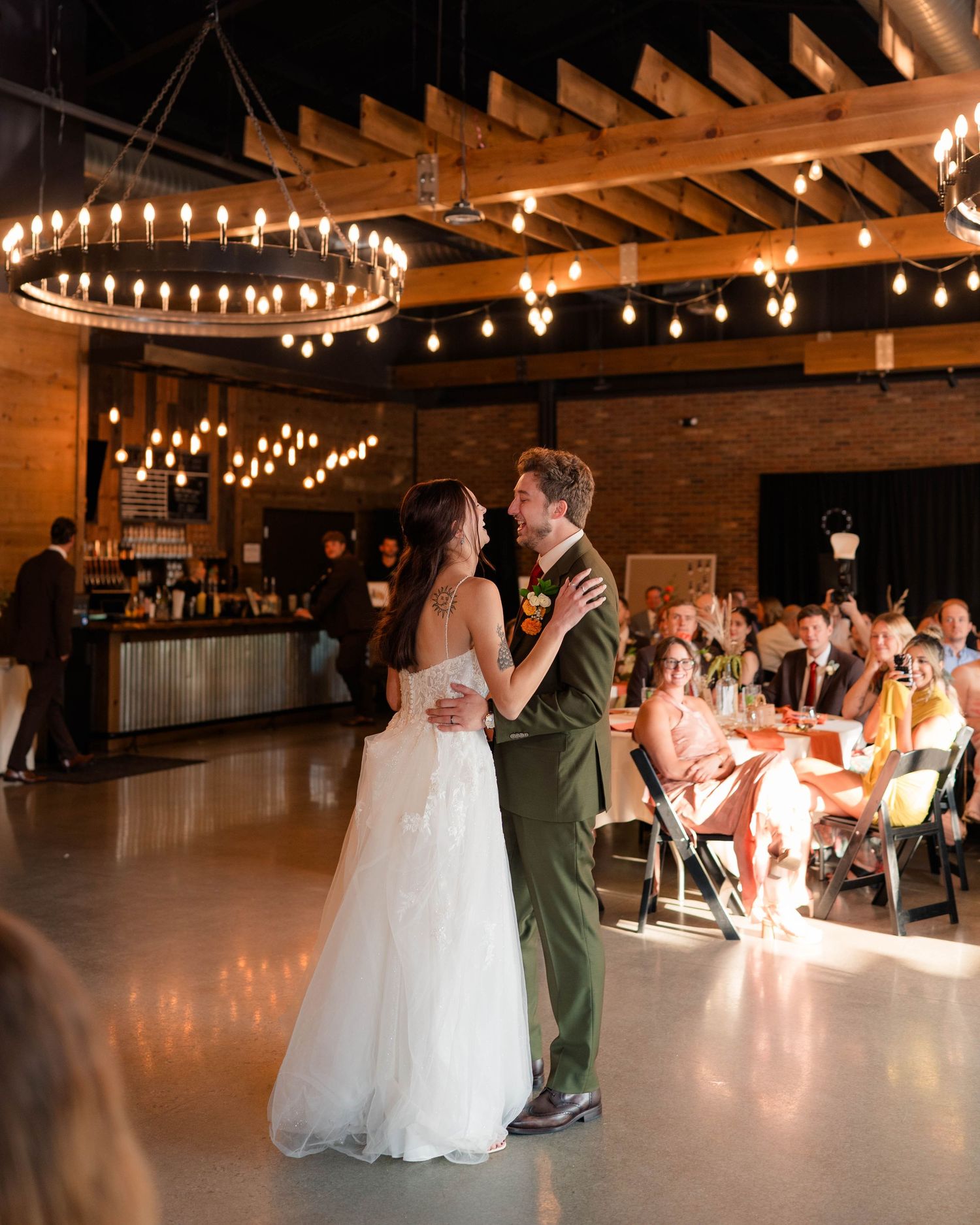 Bride and groom share first dance in rustic industrial wedding venue with warm Edison bulb chandeliers.