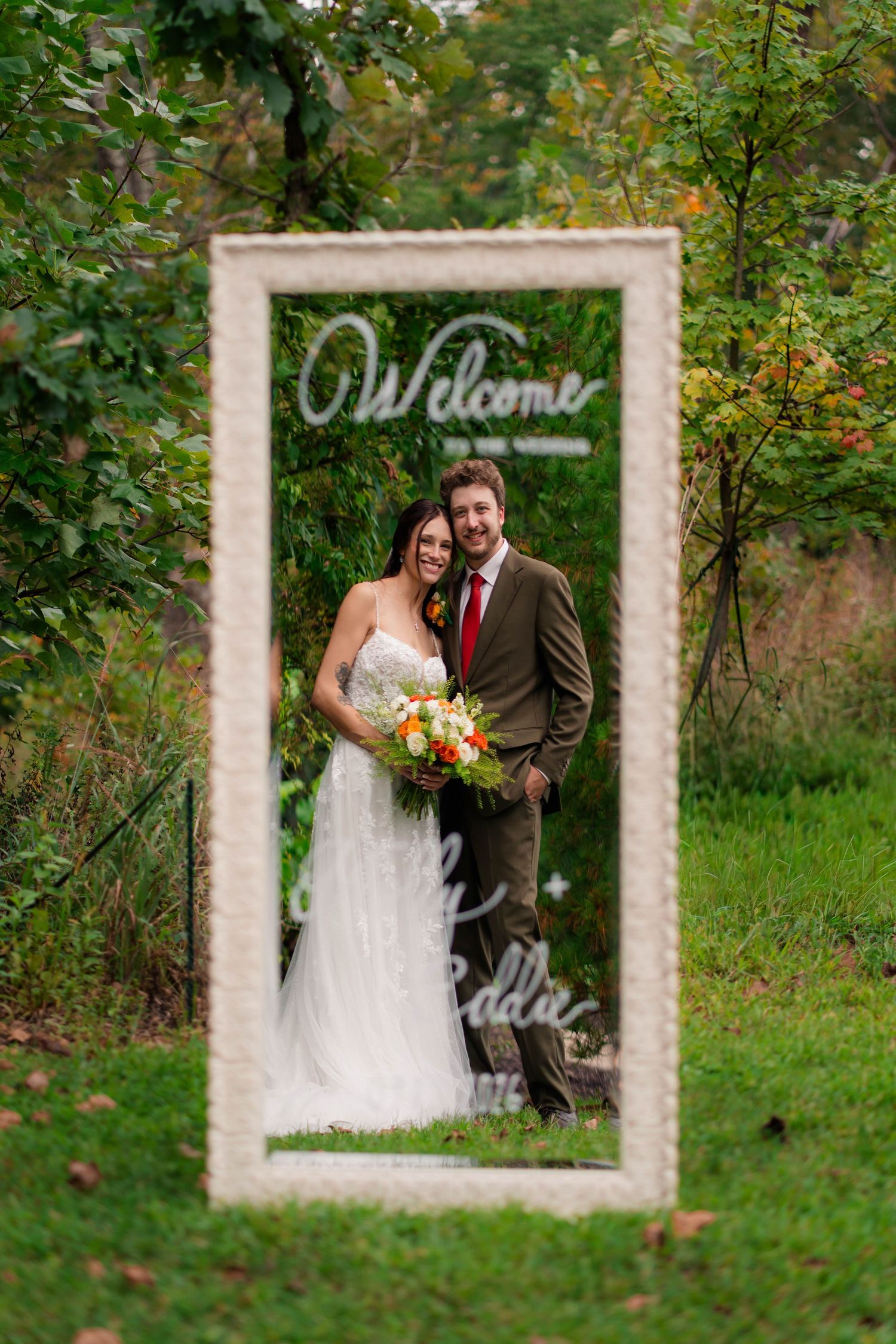 Bride and groom pose together inside a decorative welcome mirror frame at an outdoor wedding.