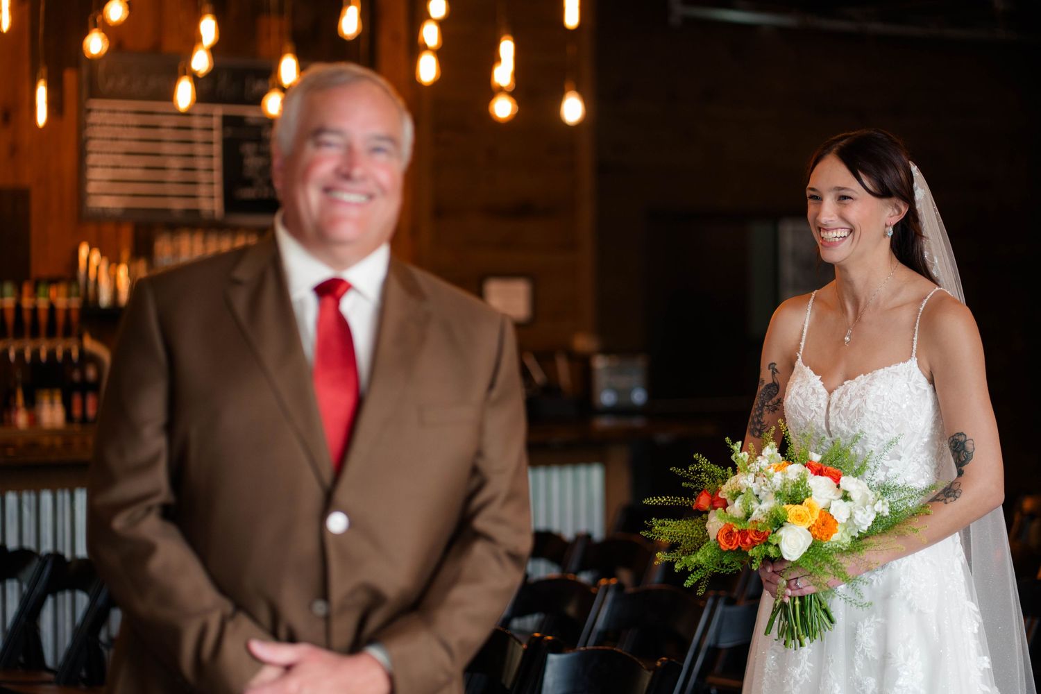 Bride in lace wedding gown smiles holding colorful bouquet during first look moment with father in brick venue.