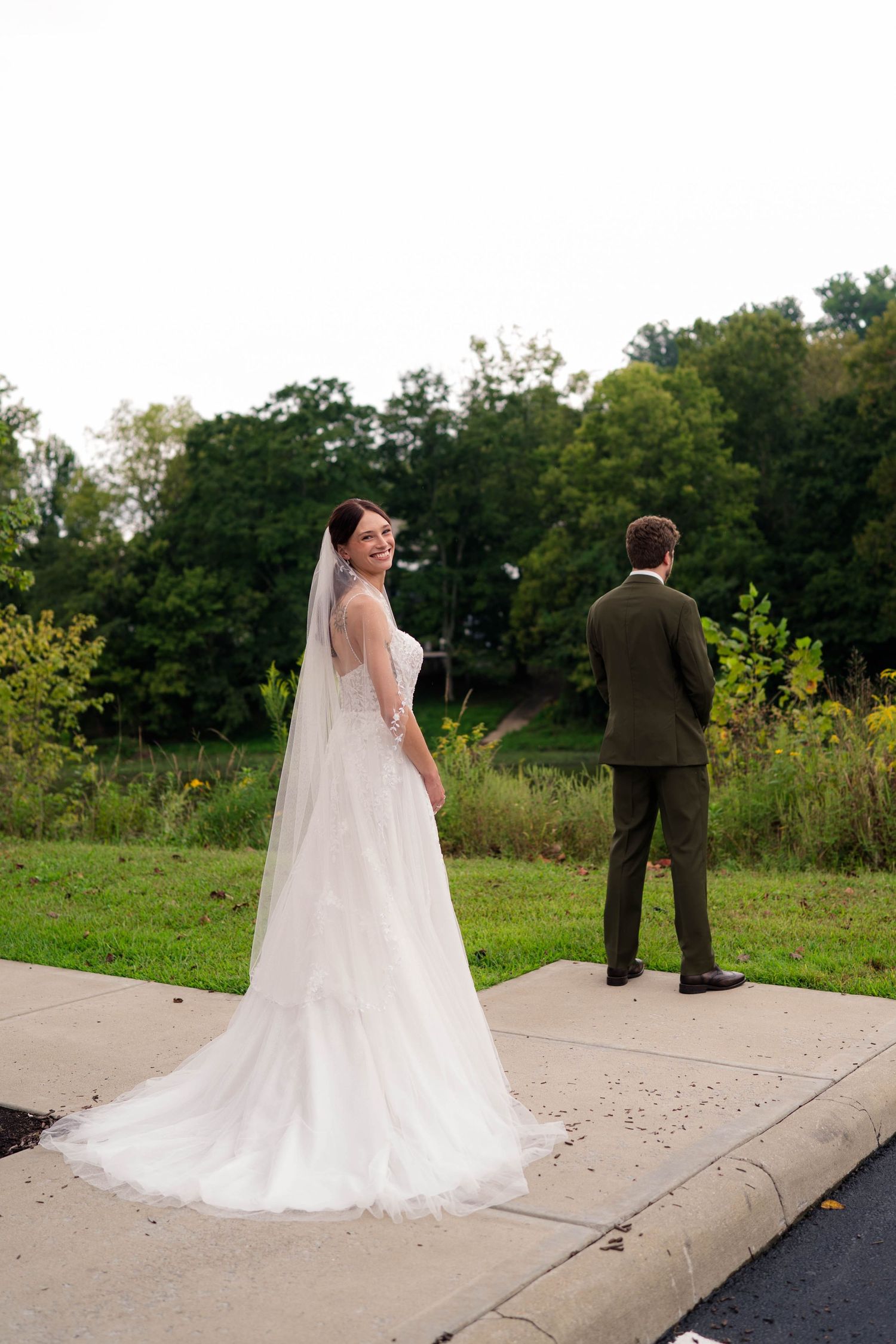 Smiling bride in white gown and veil standing beside groom in olive suit during outdoor first look.