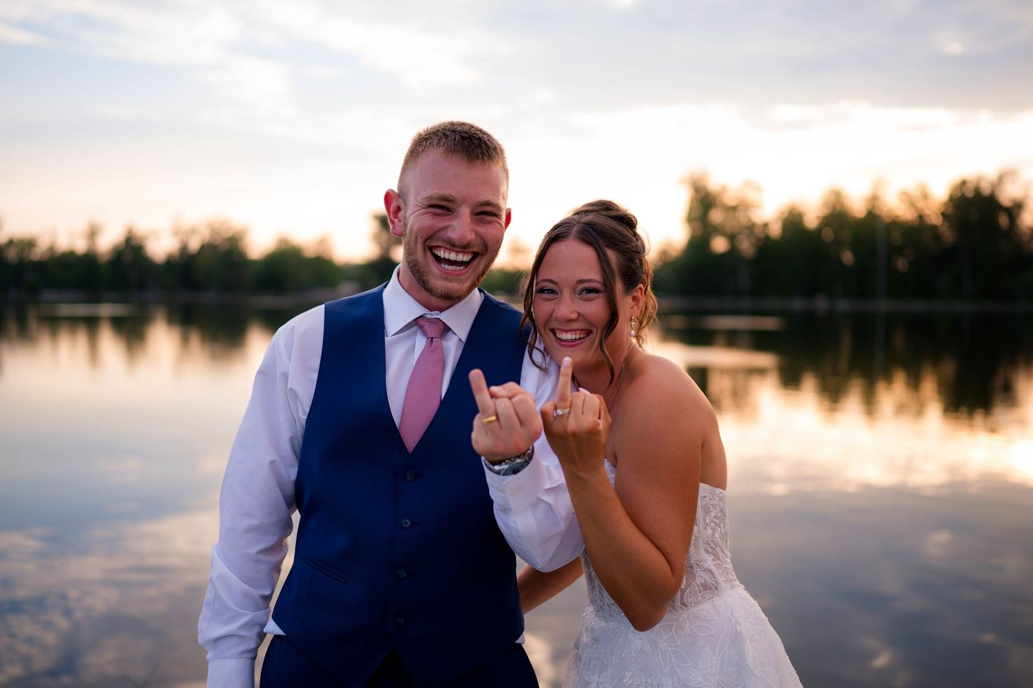 Newlyweds laugh and show off wedding rings by a lake at sunset, groom in blue vest, bride in white gown.