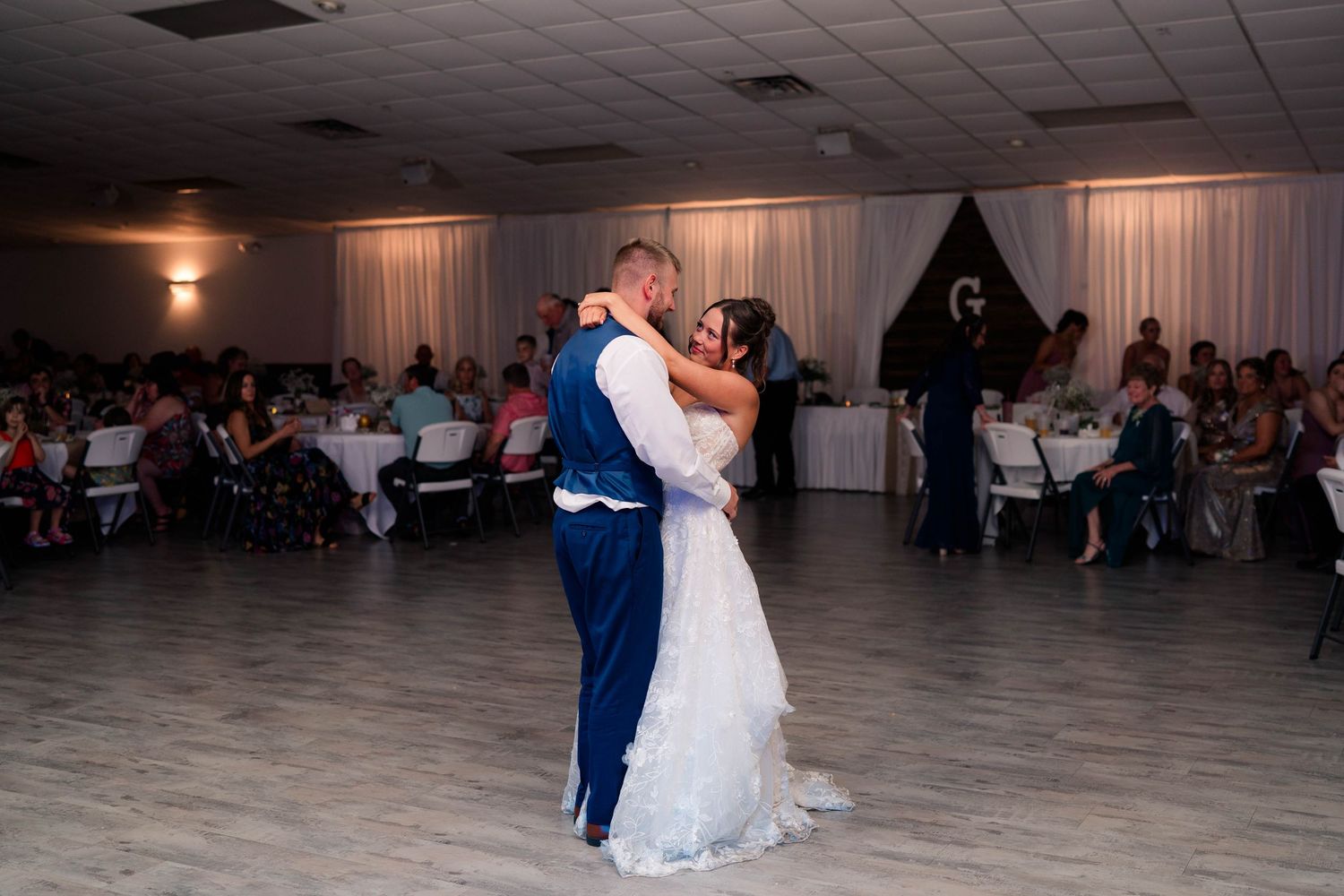 Newlyweds embrace during their first dance at wedding reception with guests watching in the background.